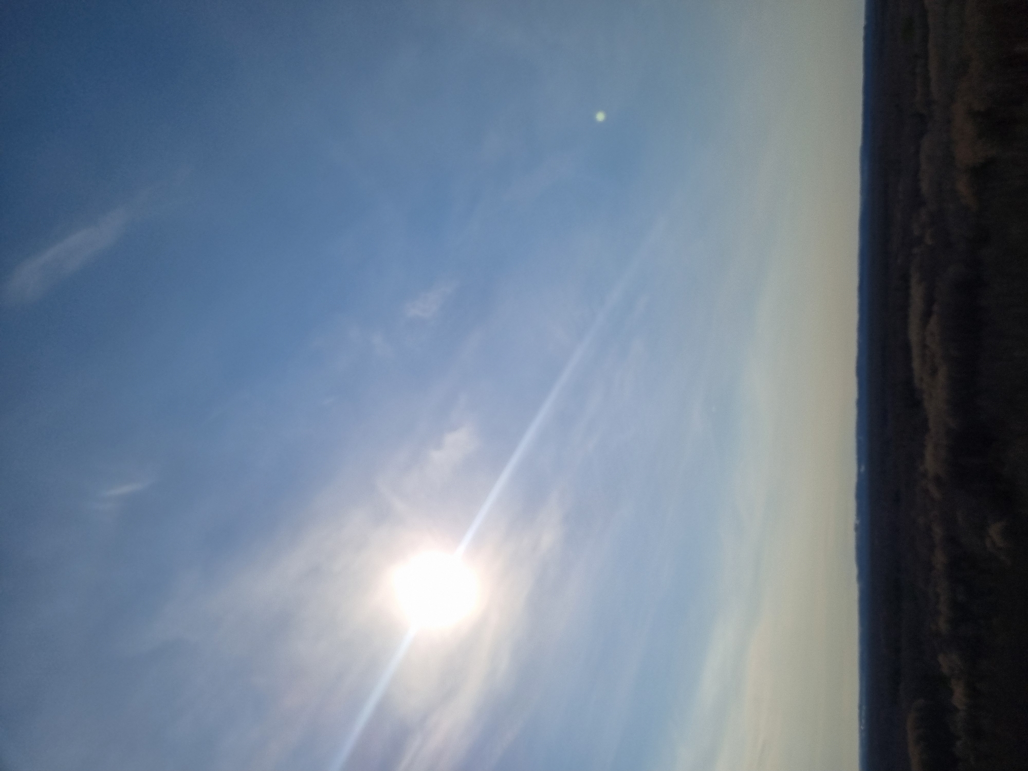 The image shows a daytime sky with a bright sun near the bottom. Wispy clouds are scattered throughout the blue sky. To the right is a strip of dark vegetation. No people or signs of urban development are visible. Given the prompt, it is assumed to be an outdoor location in Kemul, Russia, though the specific area cannot be discerned from the image.