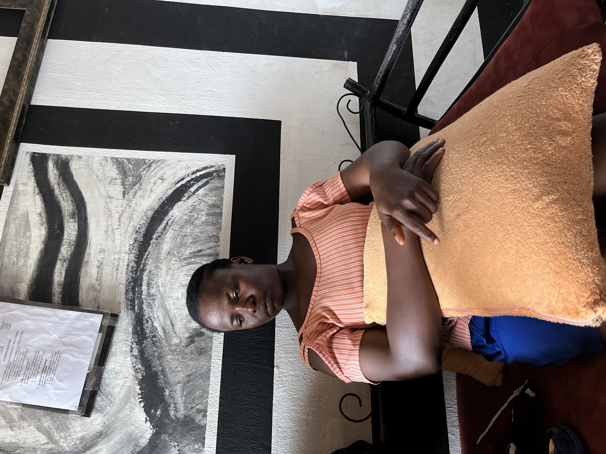 An indoor, eye-level shot captures a young woman seated in Barakin Lerawi, Nigeria. She has dark skin and short, dark hair, and wears a light orange and white striped sleeveless top. Her expression is neutral as she looks directly at the camera, her arms crossed over a large, fuzzy, light orange cushion resting on her lap. She is seated on a dark red or maroon couch or bench, with blue fabric visible beneath the cushion.
The background features a distinctive wall decorated with bold black and white horizontal and vertical stripes. To the left, an abstract artwork with swirling black, white, and grey patterns hangs on the wall. Below this artwork, a framed document displays text related to "WEDDING PACKAGES." Visible phrases from the document include "Full wedding Exp," "A comprehens," "White wedding packag," "Focused coverage for," "Traditional weddi," "edding package," and "Cultural elegance cap." A dark metal frame, possibly part of the furniture, is visible behind her. The setting suggests a reception area, lounge, or waiting room.