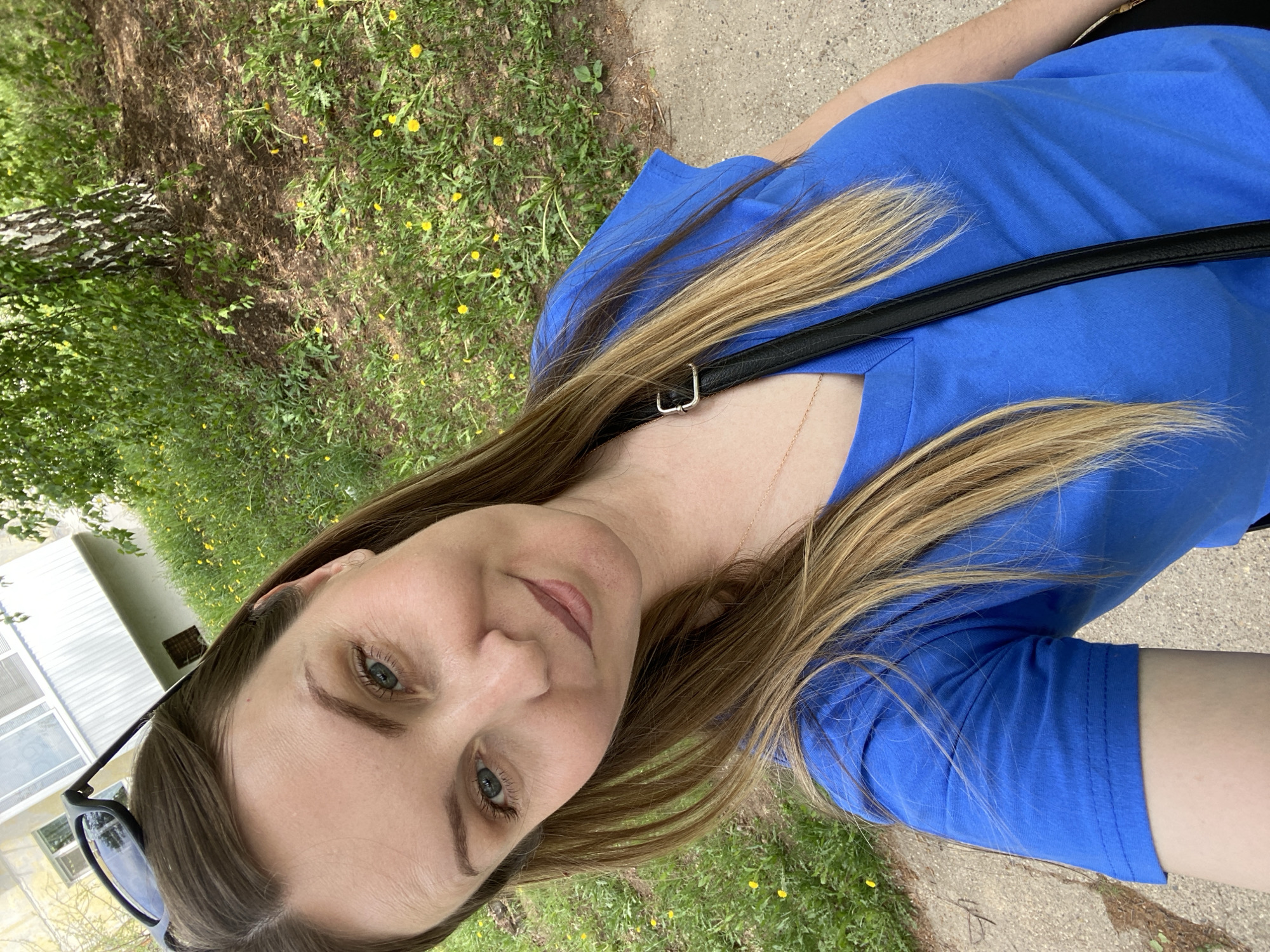 A woman is taking a selfie outdoors on a sunny day. She is wearing a bright blue t-shirt and a black shoulder bag. Her hair is long, brown, and highlighted blonde. The woman has blue eyes. She is on a sidewalk or path, next to a grassy area with some yellow flowers. In the background, there is a glimpse of a white building with windows, and some trees, including a birch tree.