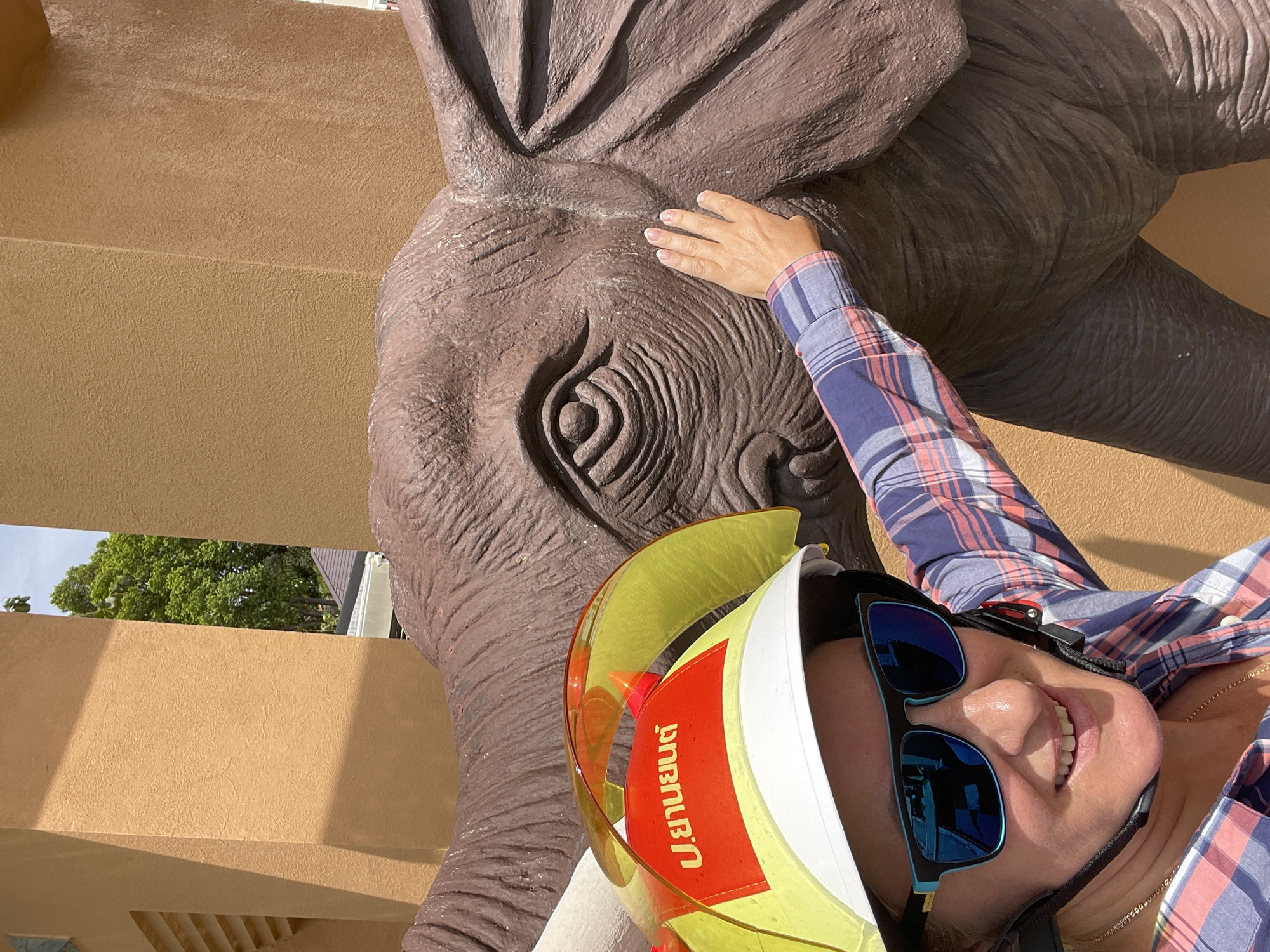 An individual wearing a yellow and white construction helmet with a clear visor and sunglasses is smiling at the camera, with their right hand touching the textured surface of a large elephant statue. The individual is wearing a blue and pink plaid shirt and a gold necklace. The elephant statue is brown and appears to be made of a rough material, showing details of its eye, ear, and trunk. The background consists of a tan building with some green trees and sky visible above. The scene is outdoors, likely in a public area or tourist attraction in Nong Pru, Thailand, indicated by the Thai writing on the individual's helmet.