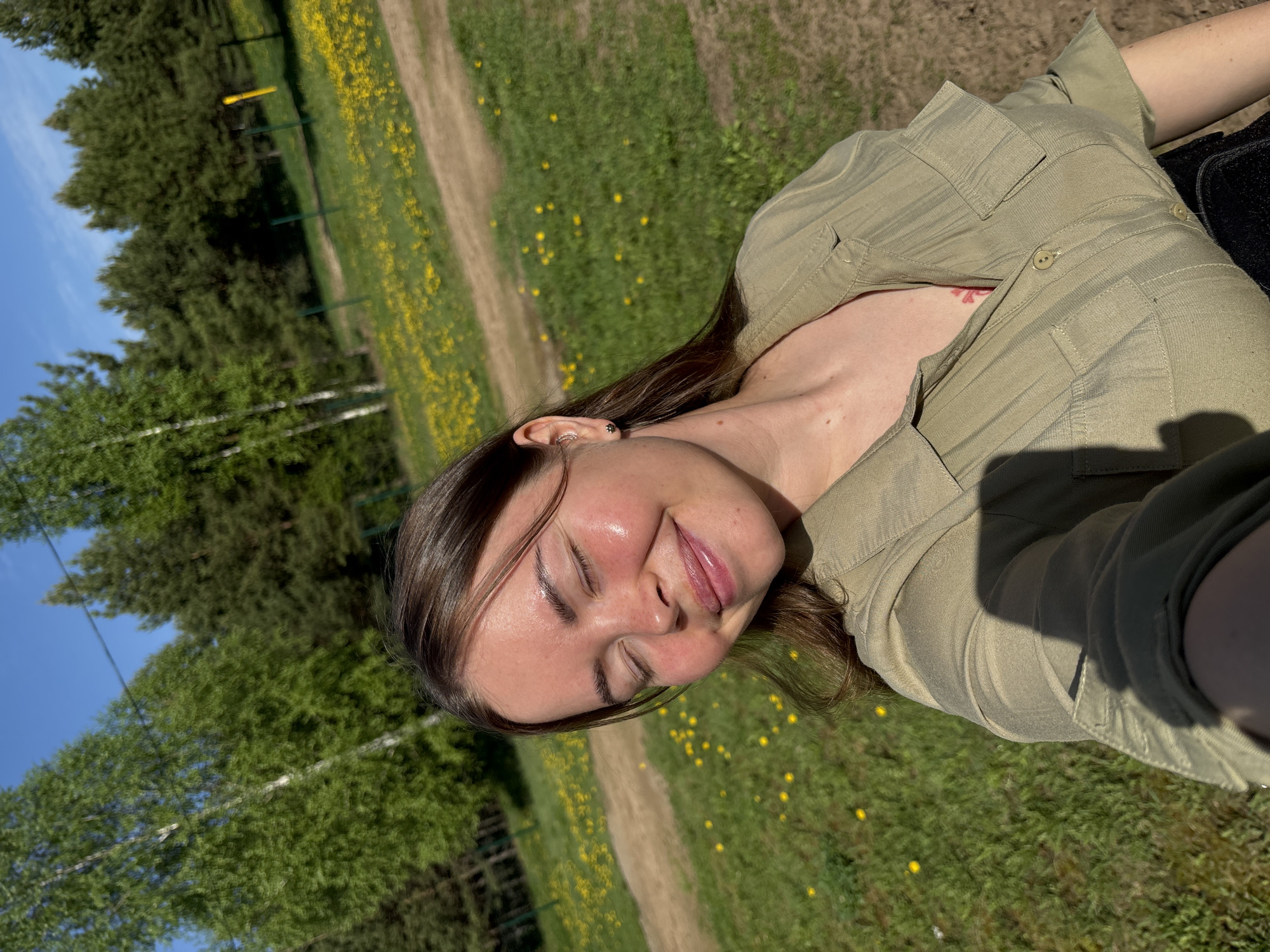 A woman is lying in a grassy area, likely in Русский Вожой, Russia. She is wearing a light green button-down shirt. She has brown hair and her eyes are closed. The background consists of green trees, a path, and green grass dotted with yellow flowers. The sky is blue in the upper left corner of the image.