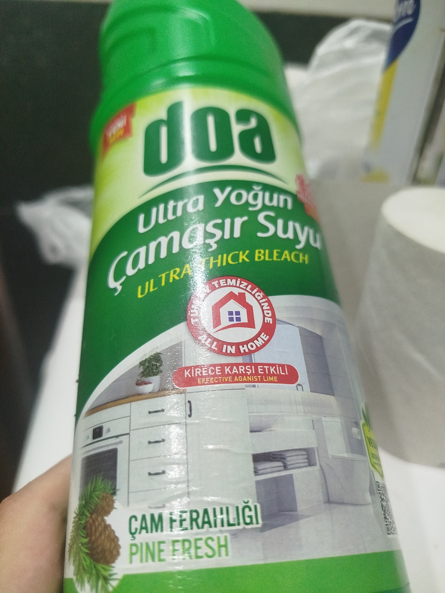 A person's hand is holding a green plastic bottle of "doa Ultra Yoğun Çamaşır Suyu" (concentrated bleach) with a "PINE FRESH" scent. The bottle has a white label with a red circular emblem depicting a house and the text "ALL IN HOME" and "TEMİZLİĞİNDE". Below this, there's a red rectangular label with text in Turkish and English, "KİREC'E KARŞI ETKİLİ EFFECTIVE AGAINST LIME". The lower part of the bottle shows an illustration of a kitchen and bathroom interior, with a pine cone and sprigs of pine needles on the left. The background is blurry and appears to be an indoor setting, possibly a store shelf or a home environment, with hints of other products and surfaces. There is no discernible information in the image to suggest a location in Batumi, Georgia.