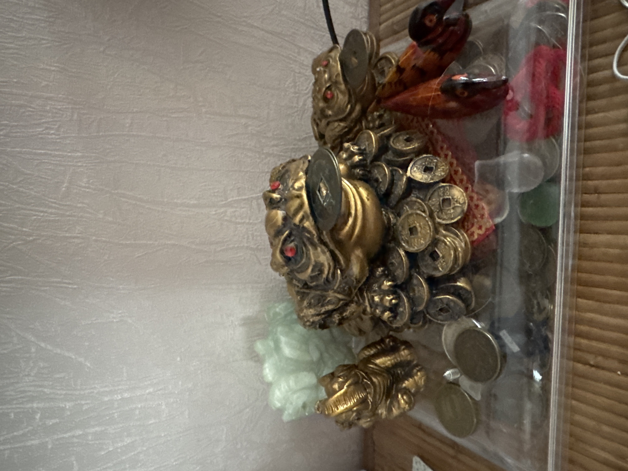 The image depicts a collection of Feng Shui prosperity frogs and other decorative items arranged on a flat surface, likely a shelf or table. In the foreground, a transparent rectangular container holds several coins of various sizes and colors, some appearing to be metallic and others possibly glass or stone. Two prominent golden frog figurines are visible. The larger frog, centrally positioned, is adorned with numerous coins and has red eyes. A smaller golden frog is placed to its left. Behind the frogs, a translucent light green object, possibly a decorative carving, is partially visible. The background features a textured wall with vertical lines, suggesting wallpaper or a similar material. The overall scene appears to be an indoor display. The provided information that this image is located in Novosibirsk, Russia, is a contextual detail that cannot be visually confirmed from the image itself.