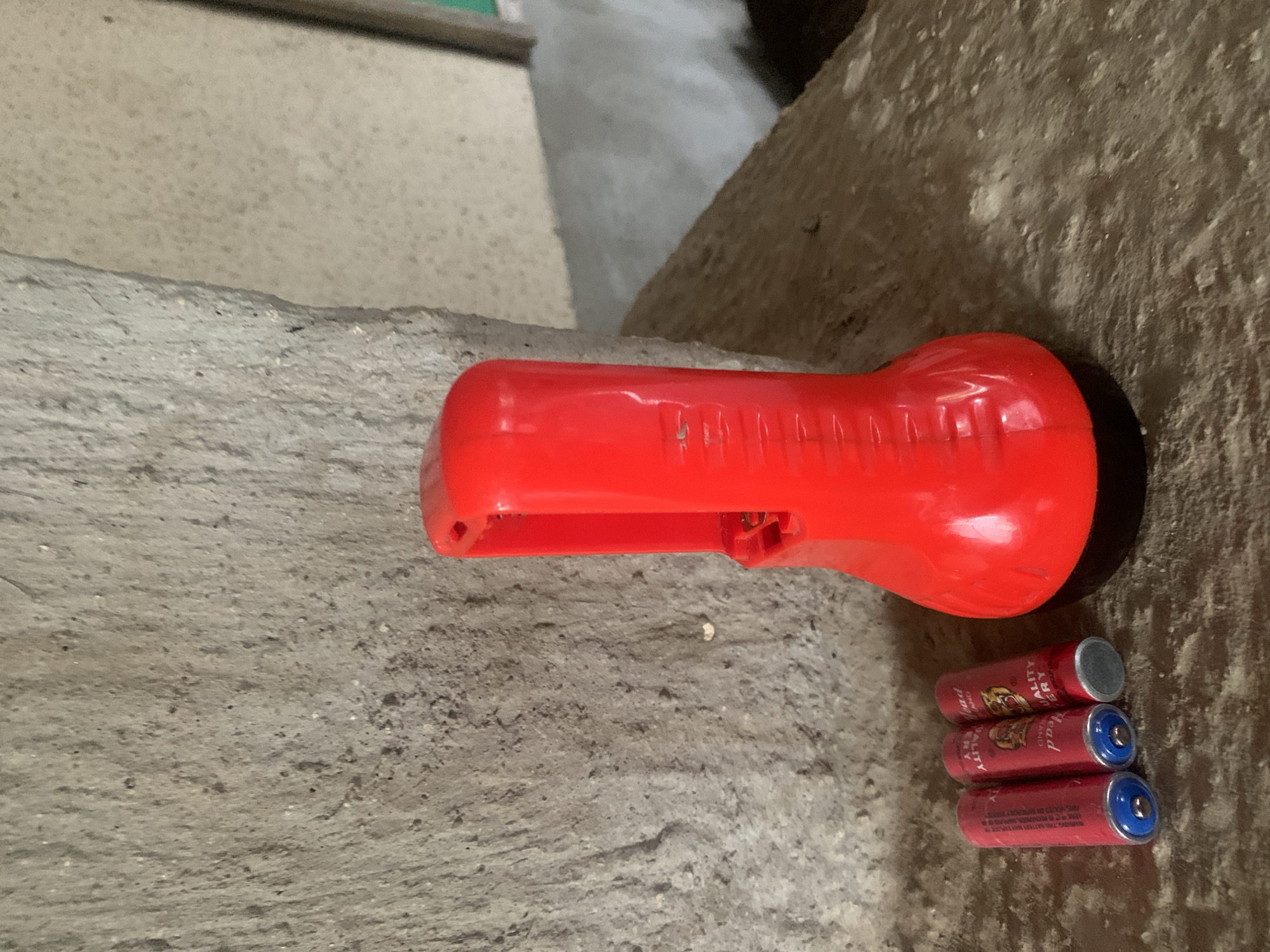 The image shows a red flashlight and three AA batteries lying on a textured surface that appears to be concrete or stone. The flashlight has a black base and a red body with a ribbed grip. The batteries are red with blue and silver ends and have branding that includes the word "HEAD" and "QUALITY BATTERY". The background is a rough, grey concrete wall. The lighting suggests an indoor or shaded outdoor environment. The scene is devoid of any human presence or activity. There is no information in the image to indicate a specific location in Jalingo, Nigeria.