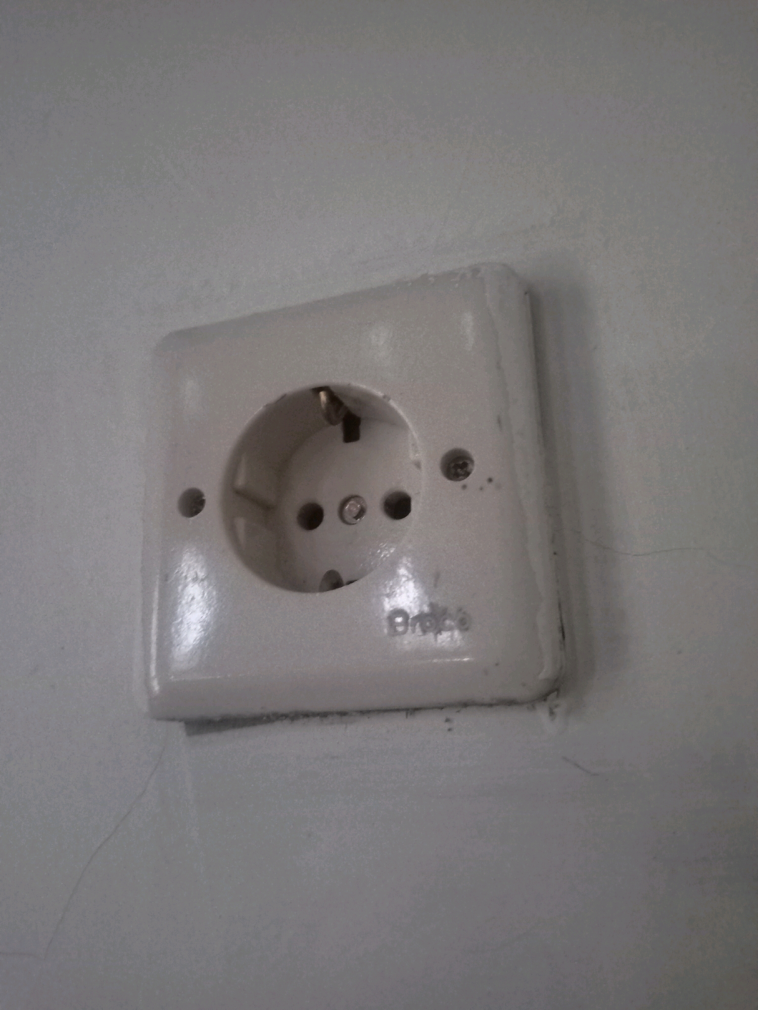 A white electrical outlet is mounted on a light gray wall in Karangtalun, Indonesia. The outlet has a rounded rectangular faceplate with a circular opening for the electrical plug. There are two small round holes above and below the main opening, characteristic of a Type E or Type F outlet. The outlet appears slightly aged, with some scuff marks and minor cracks visible on the wall surrounding it. The wall itself has a textured finish.