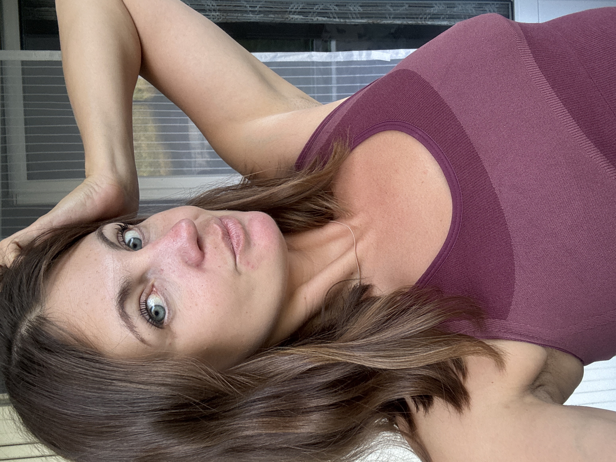 A woman lies on her side, her head resting on her arm. She wears a maroon and dark maroon ribbed tank top. Her brown hair is long and wavy, falling around her face. Her eyes are wide and blue, and she looks directly at the camera with a neutral expression. Her lips are slightly parted. The background appears to be an indoor setting with blinds or a screen visible behind her. There are no visible text elements in the image. The lighting suggests it is daytime. The overall impression is a casual, posed portrait.