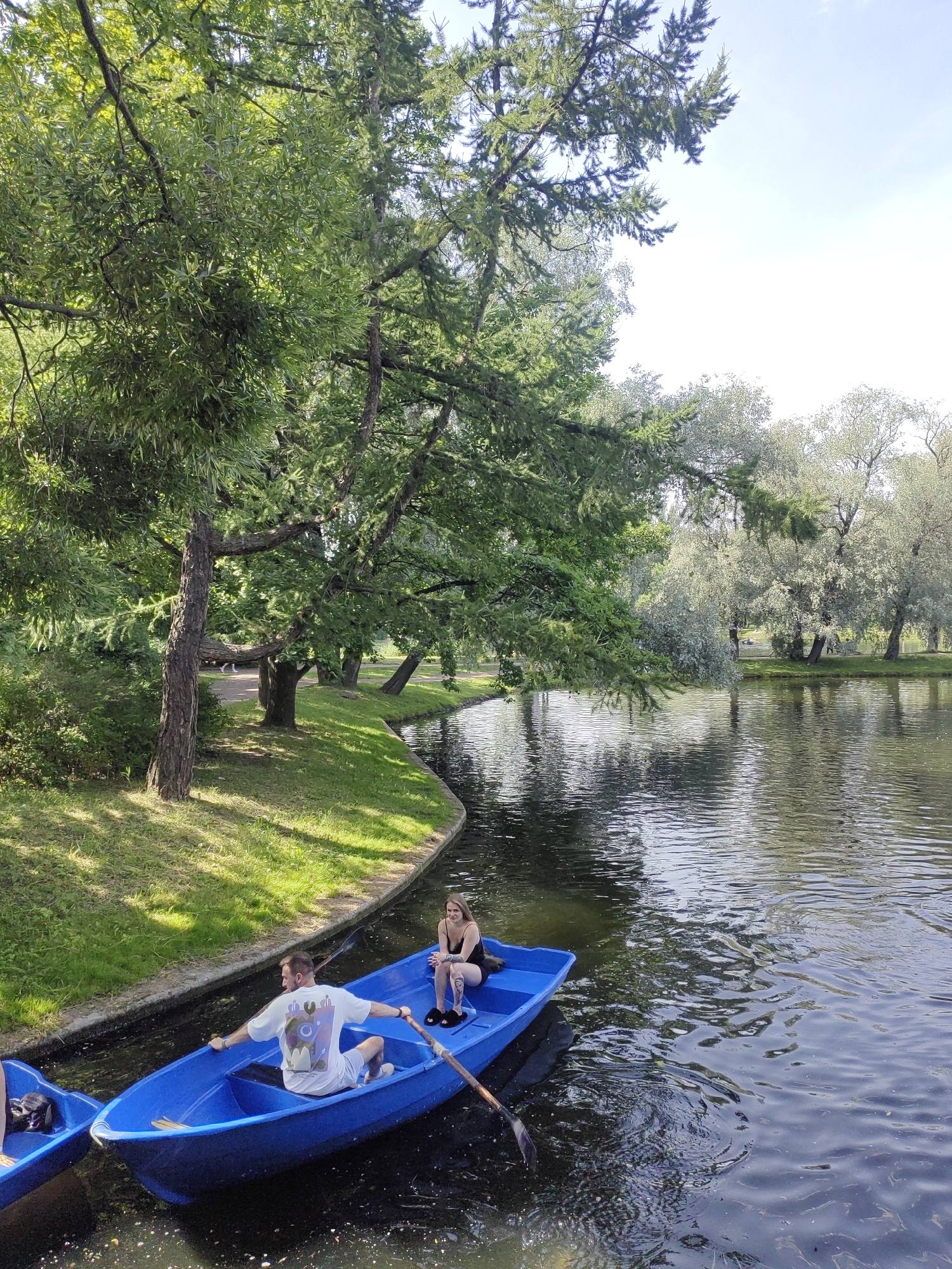 In a bright, sunlit park in Saint Petersburg, Russia, two individuals are enjoying a leisurely rowboat ride on a calm, dark body of water. The man in the front of the blue rowboat, wearing a white t-shirt and light shorts, is actively rowing with a paddle. A woman, dressed in a black top and shorts, sits in the rear of the same boat, observing the surroundings. The boat is positioned in the foreground, with its bow pointing towards the left side of the frame. A second blue rowboat is partially visible in the lower left corner. The park features lush green grass lining the water's edge, with a variety of trees providing ample shade. The water's surface reflects the sky and the surrounding foliage, creating a dappled, serene atmosphere. The background reveals more trees and a clear blue sky with a few wispy clouds.