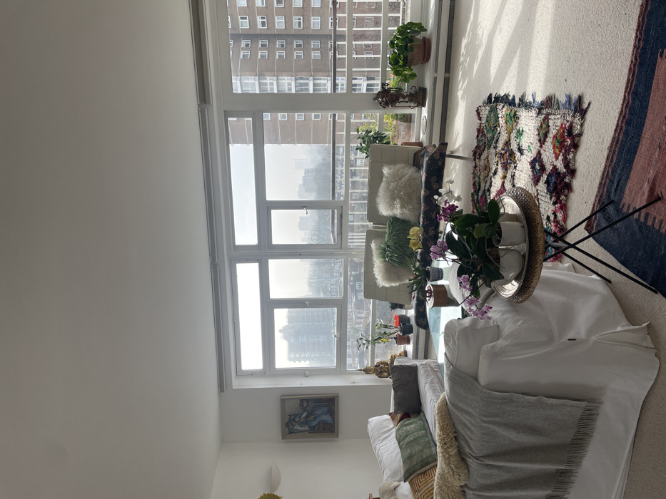 The image depicts the interior of a brightly lit living room located in a multi-story residential building in London, United Kingdom. The room is furnished with a white slipcovered sofa on the left, adorned with multiple throw pillows in shades of green, brown, and cream, and a grey blanket. To the right, two white lounge chairs with white frames are visible, each featuring a large, fluffy cushion—one green, one white. A clear glass coffee table stands between the sofa and the lounge chairs.
In the foreground, a small, round, ornate metallic side table with three dark legs holds a silver tray containing three white potted orchids with purple and yellow flowers, alongside two smaller white pots. The floor is covered by light-colored carpeting, over which two distinct rugs are placed: one colorful, shaggy rug with various geometric patterns in shades of white, red, blue, and green, and another rectangular rug in blue and pink tones.
The rear wall features a large, multi-pane window spanning the width of the room, offering a view of a city landscape under a bright, somewhat hazy sky. Through the window, several other residential buildings are visible, including a prominent reddish-brown brick high-rise with numerous windows on the right. Trees without foliage are visible in the mid-ground, suggesting a colder season. A metal balcony railing is present immediately outside the window. The windowsill displays a collection of potted plants, a golden Buddha statue, and an ornate, dark metal sculpture. On the left wall, a framed painting with blue and brown tones is hung. No individuals are present, and no active interactions are occurring within the scene.