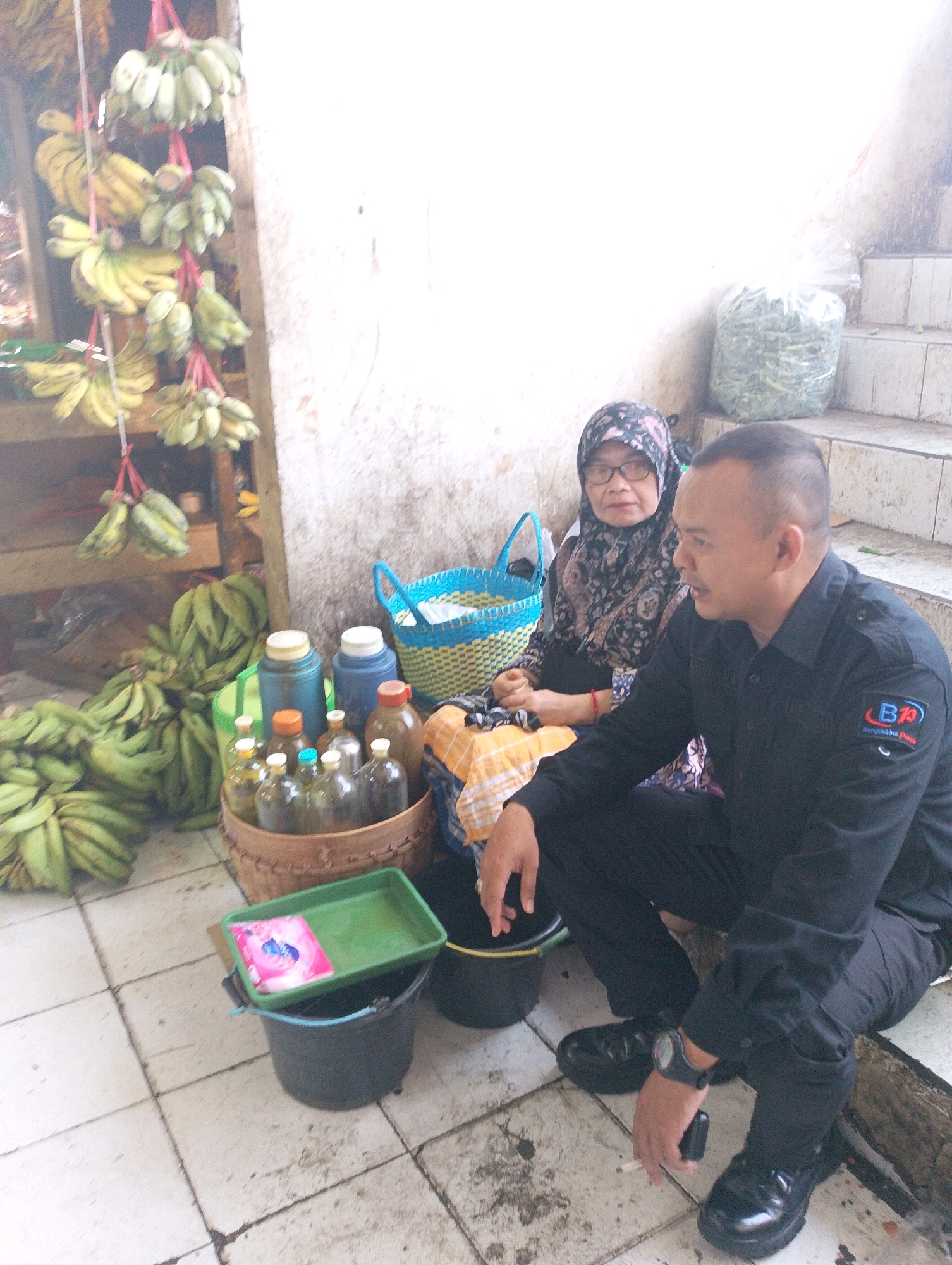A medium shot captures a street scene in Lembang, Indonesia, where two individuals are seated on the ground amidst a collection of produce and bottled goods. In the foreground to the right, a man in a black collared shirt and dark pants is seen from the chest down, looking to his left. He has a watch on his left wrist and is holding a cigarette in his right hand. To his left, a woman wearing a headscarf and patterned shirt is also seated, facing the viewer. In the background, several bunches of green bananas are displayed on a wooden shelf and also piled on the ground. Various bottles, some containing liquids, are arranged in baskets and on the ground, alongside blue and yellow woven baskets. A large plastic bag filled with green produce is visible further in the background, near a set of stairs. The scene is set against a textured wall and a tiled floor.