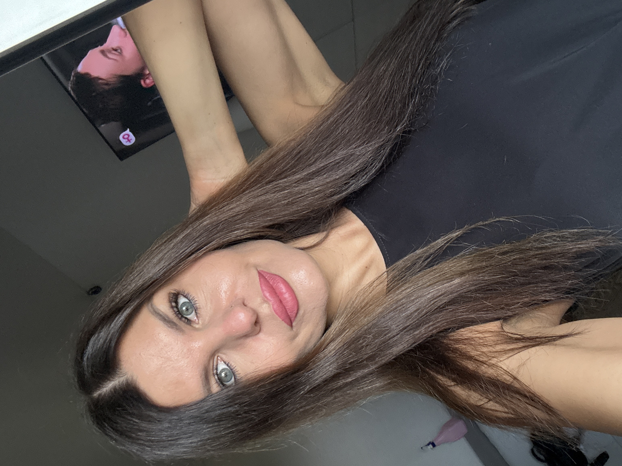 A woman with long brown hair and blue eyes is taking a selfie. She is wearing a black top. Her left arm is raised and her hand is behind her head. In the background, a television screen displays a profile of a person with a speech bubble containing a "+0" symbol. The scene appears to be indoors, in what could be a residential setting. There is no information available to determine the specific location in the city of Chaikovsky, Russia.