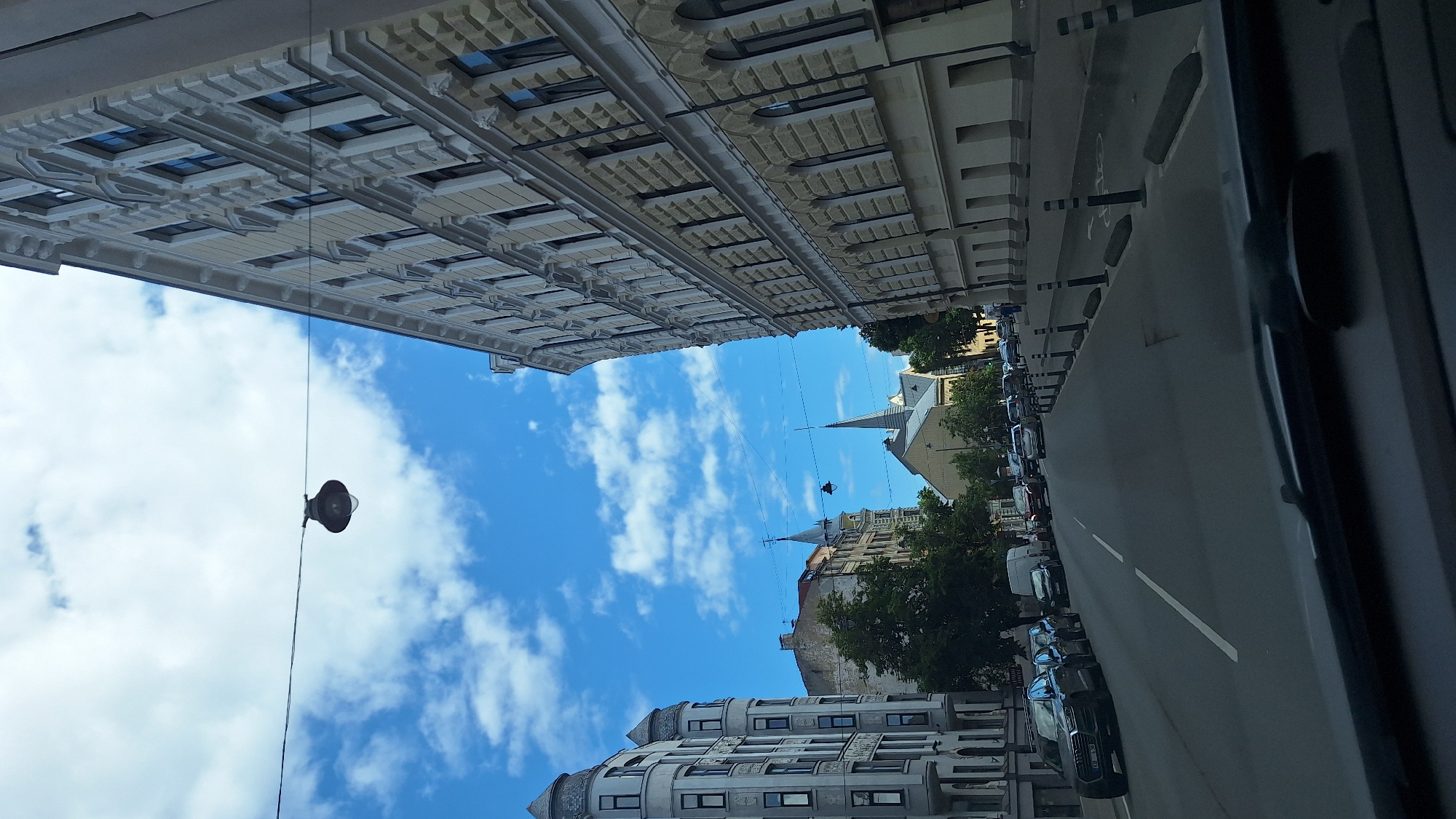 The image shows a street scene in Riga, Latvia, on a partly cloudy day. The perspective is from inside a vehicle, looking forward. The street is lined with multi-story buildings on both sides, featuring ornate architectural details and balconies. On the left side of the street, there are several cars parked, including a dark grey Audi SUV. On the right side, the street continues with more buildings and a sidewalk with bollards.
Further down the street, there are more vehicles, including vans and cars, moving or parked. In the distance, church spires and rooftops of older buildings are visible, indicative of an urban environment with historical architecture. The sky is blue with scattered white clouds. A street lamp hangs from wires stretched across the street in the upper left portion of the image. The overall impression is a typical European city street.