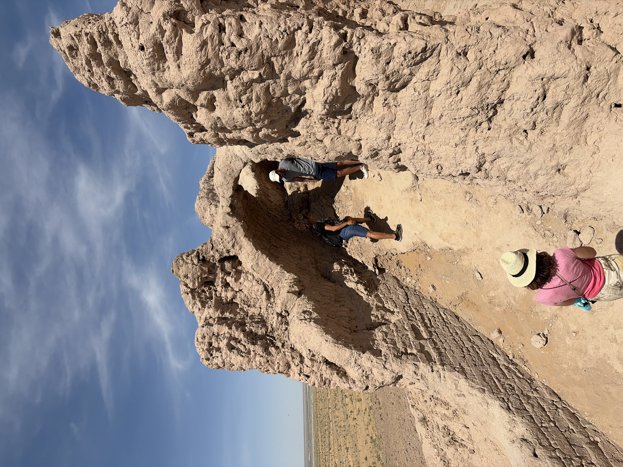 The scene is set in ancient, eroded mud-brick or adobe ruins under a clear blue sky with sparse, wispy white clouds, in the city of Unknown, Uzbekistan. The structures are of a light brown/tan color, exhibiting rough, textured surfaces, crumbling sections, and visible layers suggesting construction from stacked blocks or packed earth. An arched passageway is prominent in the midground, with towering, angular masses forming parts of the ruins, likely remnants of a fortress or large settlement. In the distance, a flat, arid desert landscape with sparse, low-lying scrub vegetation extends to the horizon.
Three individuals are present, appearing to explore the ruins. In the immediate foreground, with her back to the camera, is a female wearing a light-colored straw hat with a black band, a pink short-sleeved top, and light-colored patterned shorts. A light blue item hangs from a strap across her body. She is positioned on a downward-sloping path within the ruins, looking ahead.
Further into the scene, beneath the main arched section, are two males. The man on the left wears a dark t-shirt, blue denim shorts, black sneakers, and a black backpack. He is looking downwards and slightly to his right, partially obscured by shadow. The man on the right wears a light grey t-shirt, dark blue shorts, a white baseball cap, and white sneakers. He is standing slightly to the right and a step above the first man, also looking downwards and to his left, seemingly engaging with the ground or the other individual. The ground within the ruins is sandy and uneven, with scattered small rocks and debris.