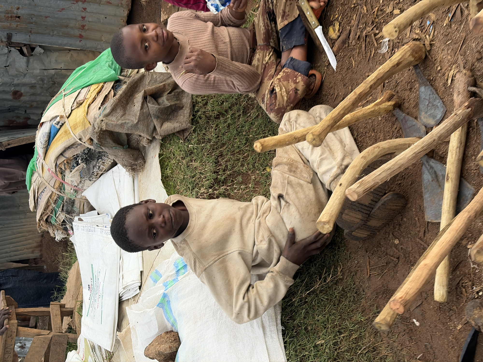 In Gura Top, Nigeria, two young boys are seated on the ground amidst a collection of agricultural tools and supplies. The boy on the left, dressed in a light brown sweatshirt and pants, appears to be contemplating something as he rests his hand on his leg. To his right, another boy, wearing a light brown ribbed sweater and patterned pants, is looking towards the camera with a slight smile and holding something in his raised hand. Scattered around them are several wooden tools, likely for farming, with metal blades visible among them. In the background, there are piles of sacks, some white with green lettering, and other bundled materials covered with a green sheet. A corrugated metal wall forms the backdrop, suggesting a rustic or market-like environment. The ground is a mix of dirt and sparse grass.