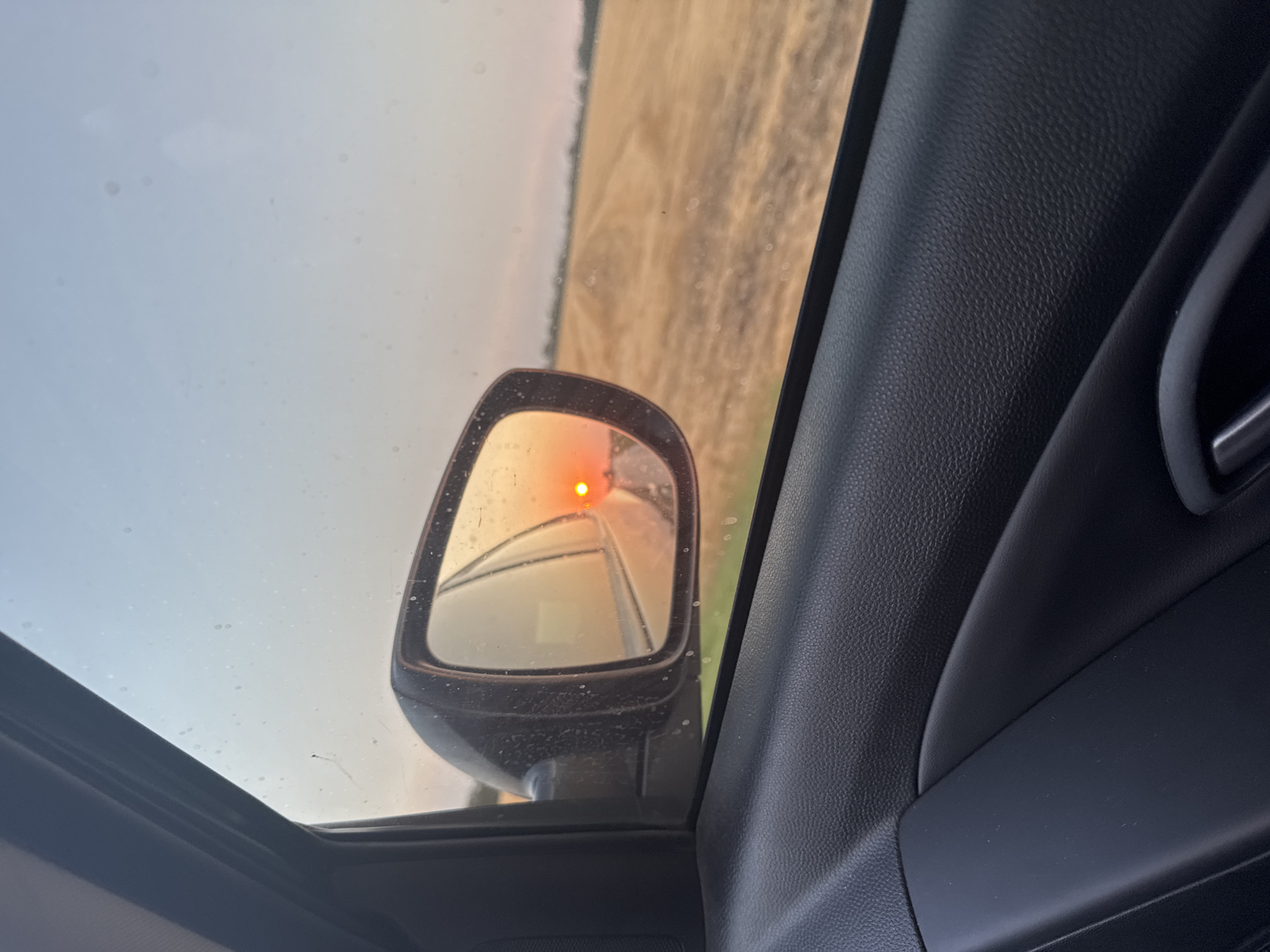 The image captures a sunset viewed from inside a vehicle, looking out the passenger side window and into the side mirror. The side mirror reflects a bright orange sun low on the horizon, casting a warm glow over a harvested field of golden stubble. The field stretches towards a distant treeline under a hazy, pale blue sky. The interior of the car, including the black textured door panel and armrest, is visible in the foreground. The image is taken from a roadside perspective, suggesting a journey through a rural landscape. The location is identified as Magistralny, Russia.