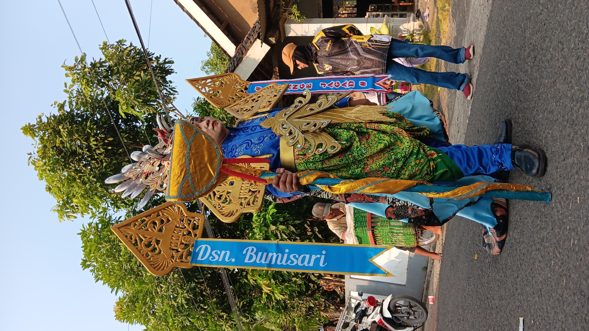 In the city of Cerme, Indonesia, a person dressed in a vibrant, ornate costume stands on a paved road. The costume features a blue shirt with gold embroidery, a golden belt, and a green patterned garment draped over the front. Intricate golden headdresses and shoulder pieces adorn the individual, resembling stylized wings and mythical creatures. The person holds a long, colorful staff.
Behind the main subject, a blue banner with white lettering reading "Dsn. Burnisari" is visible, suggesting the name of a hamlet or village. Other individuals are present in the background. To the left, a man in a cap is partially obscured behind a woven green structure, possibly a cart. To the right, a person wearing a black and yellow shirt with the logo "RWDI" and jeans stands with their hands behind their back. A motorcycle is parked on the left side of the road. The scene is set outdoors, with trees and a building with a tiled roof in the background, under a clear sky. The ground is asphalt.