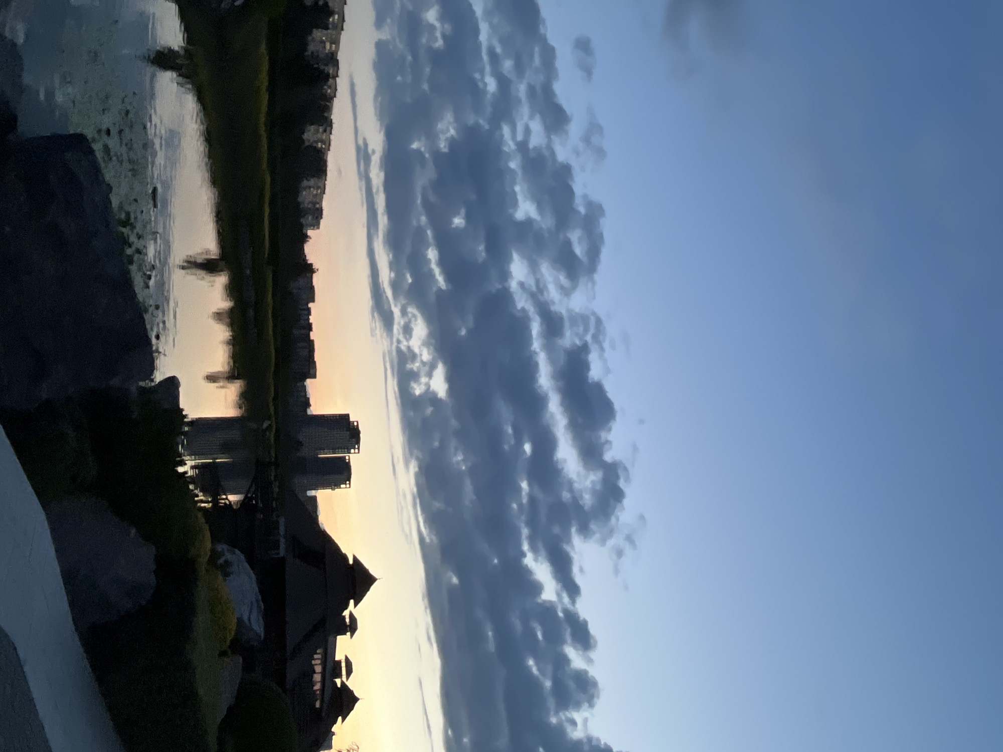 This is a vertically oriented, low-angle photograph taken at dusk in Dnipro, Ukraine. The scene depicts a tranquil waterside setting with urban elements.
In the foreground, to the left, a paved walkway runs along the edge of a body of water. Large rocks and dark green shrubbery are visible along the embankment. A dark, multi-peaked building, possibly a pavilion or unique architectural structure, dominates the lower right portion of the frame.
The middle ground reveals a calm body of water, reflecting the sky and the buildings on the opposite shore. The reflections show several tall, modern buildings, appearing as silhouetted towers against the fading light. A thin line of green vegetation separates the water from the buildings.
The background is dominated by the sky, which is a gradient of soft blue transitioning to a warm, pale yellow near the horizon. A prominent band of dark, textured clouds stretches across the sky from left to right, adding dramatic contrast to the serene setting. The overall mood is peaceful and contemplative, characteristic of a sunset or twilight hour. There is no visible text in the image.