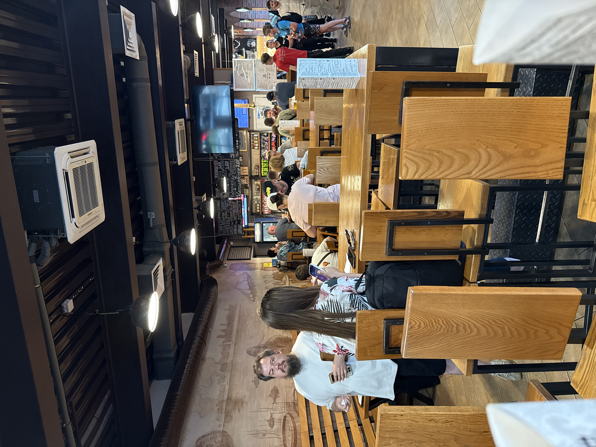 The image depicts the interior of a casual dining establishment, possibly a pub or restaurant, with wooden tables and chairs. In the foreground, empty wooden chairs are visible, some with backs turned towards the viewer. The midground shows a man with a beard and a white t-shirt, holding a smartphone, standing next to a woman with long brown hair wearing a floral-patterned top. They are seated at a wooden table. Further into the establishment, other patrons are seated at tables, some engaged in conversation, while others appear to be waiting. There are also shelves with bottles and menus visible in the background, as well as television screens mounted on the wall. The lighting is ambient, with overhead lights illuminating the space. The overall atmosphere suggests a busy, social gathering place. The presence of the mural on the wall, depicting a historical scene with boats and buildings, might indicate a themed establishment. Given the context of a query about Samara, Russia, this establishment could be located in that city.