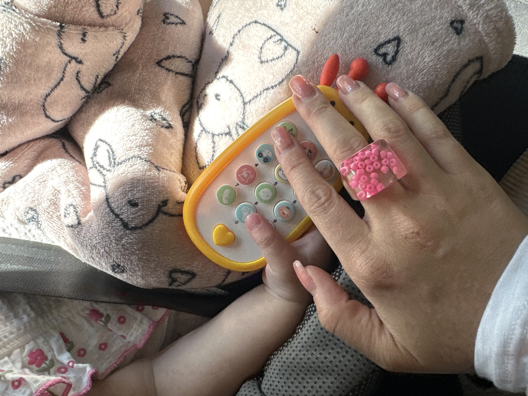 This image captures a close-up, top-down perspective of an adult and a child interacting with a toy, likely situated in Nizhny Novgorod, Russia. The scene is brightly lit by natural sunlight, suggesting it's daytime.
The main subjects are an adult's hand and a child's arm and hand. The adult's hand is positioned over a yellow and white toy phone or remote control. This hand features long, light pink manicured nails and wears a distinctive clear, square ring filled with numerous tiny pink beads. The child's left arm and hand are visible reaching towards the toy, with a finger seemingly touching one of its buttons.
The toy itself is a vibrant, triangular-shaped device with a yellow border and a white central panel featuring several round, colorful buttons. These buttons display various icons including a car, a cloud, an apple, a smiley face, a star, and numerical digits, along with a prominent yellow heart-shaped button.
The background consists primarily of a soft, fluffy pink blanket or fabric adorned with black outline drawings of what appear to be deer or bunnies, some accompanied by small heart shapes. A dark, textured fabric, possibly part of a car seat or carrier, is visible beneath the blanket and toy. A small section of white child's clothing with pink floral and dotted patterns can also be seen. The adult's white sleeve is visible on the right side of the frame. No specific text is readable, only icons and numbers on the toy buttons. The image conveys a tender moment of play and interaction.