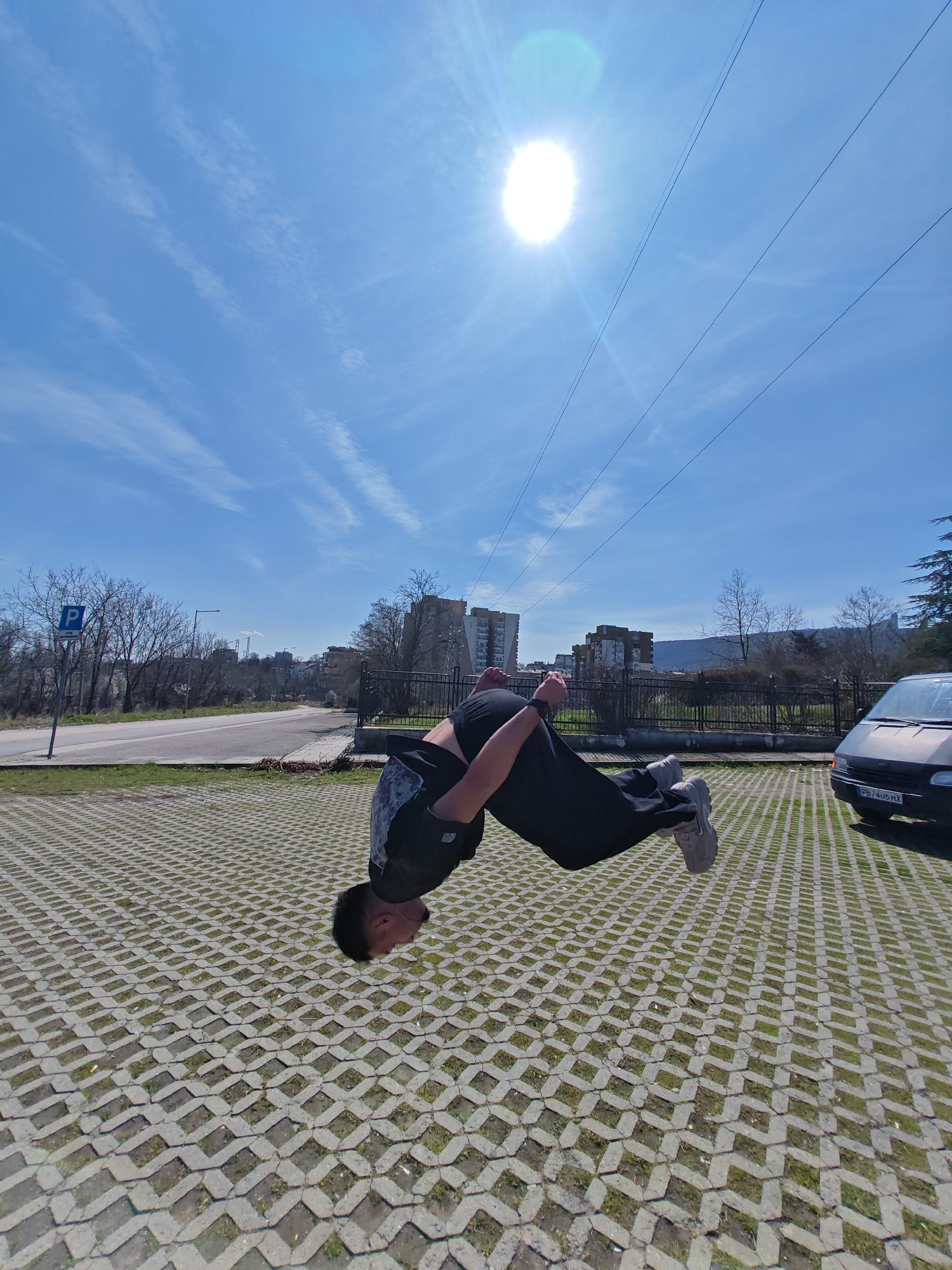 An outdoor, daytime image captures a male individual performing a backflip or similar acrobatic maneuver, suspended mid-air in an inverted position. The individual, clad in a dark short-sleeved top with a faint pattern on the back, dark athletic pants, and light grey sneakers, has his head tucked towards his chest and arms bent.
The ground surface consists of a concrete grid paver system with grass growing within the interstices. To the left, a paved road curves away from the foreground, bordered by a grassy verge and a blue "P" parking sign on a pole. Bare deciduous trees are visible along the road. To the right, a dark-colored van, bearing the Bulgarian license plate "PB 7405 HX", is parked on the same paved surface. A black metal fence runs horizontally across the midground, separating the paved area from an overgrown embankment and additional bare trees.
In the background, multiple multi-story residential buildings are visible, constructed from concrete or light-colored materials. Behind these structures and trees, a prominent, light-colored rocky massif, identifiable as the Shumen Plateau, rises under a clear blue sky. Thin, dark utility lines traverse the upper portion of the sky, where the bright sun creates a distinct lens flare effect. The scene is situated in Shumen, Bulgaria, specifically within an urban or suburban parking area adjacent to a thoroughfare, with residential complexes and the natural landmark of the Shumen Plateau in the distance. The bare trees indicate late winter or early spring.