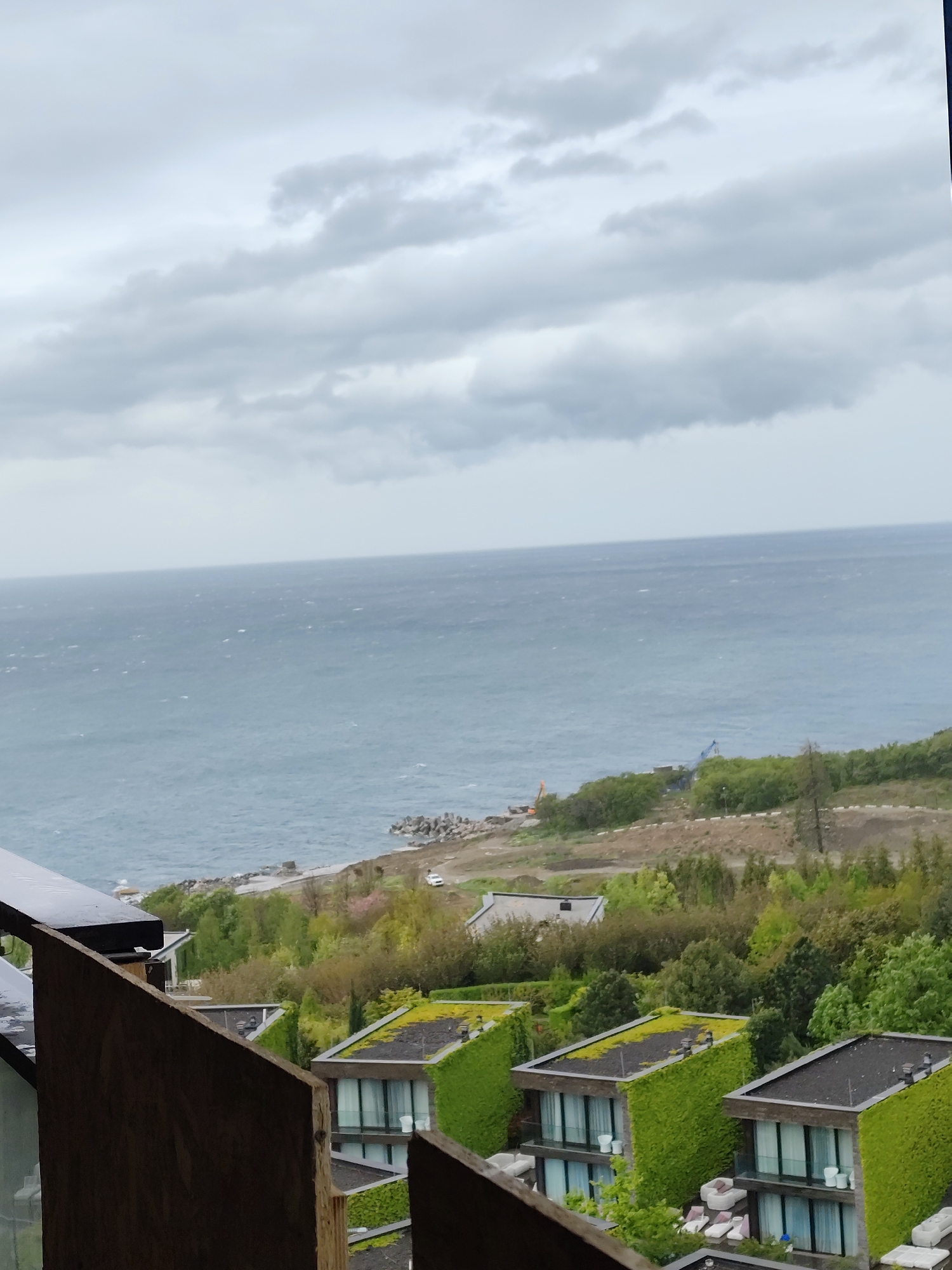 The image captures a view of the sea and coastline near Симеизский поселковый совет, Ukraine, under an overcast sky. Several modern buildings with green roofs and ivy-covered walls are visible in the foreground. In the midground, there's vegetation and a road. In the background, the sea meets the horizon, and construction equipment is visible on the coastline.