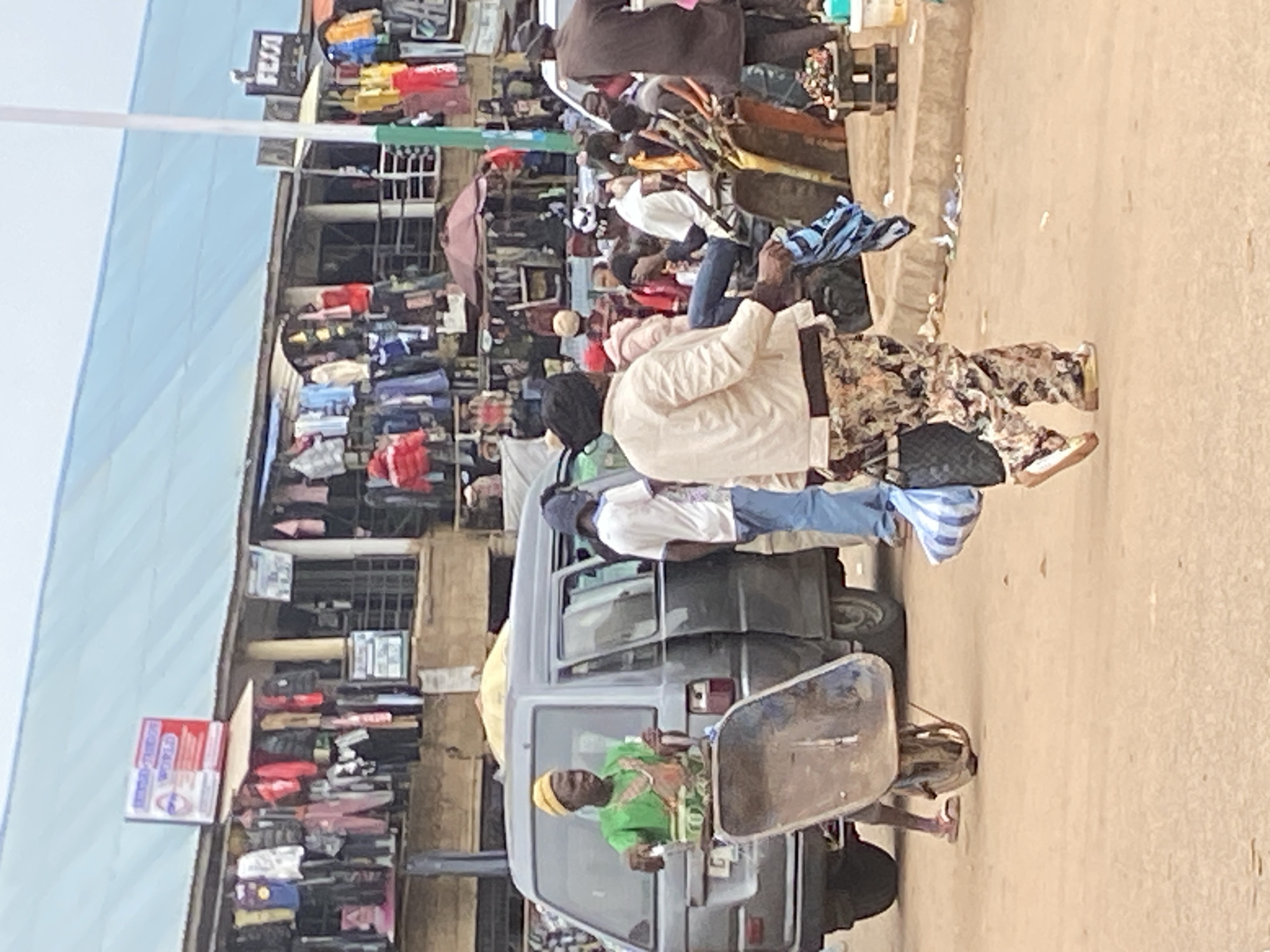 The image depicts a bustling market scene in Bukuru, Nigeria. Several people are visible, with a primary focus on individuals interacting within the market stalls and near a van. The market appears to be an outdoor setting, with stalls overflowing with various goods, predominantly clothing and textiles, displayed on shelves and racks.
One person, wearing a light-colored jacket and patterned pants, is prominently positioned in the foreground. Another individual in a green top and yellow head covering is seated in the driver's seat of a grey van, which is parked amongst the stalls. Other individuals are seen browsing or walking through the market.
The activity suggests a typical day of commerce, with people engaged in shopping and selling. The time of day appears to be daytime, likely morning or afternoon, given the ambient light. The weather seems fair, contributing to the outdoor market activity.
Notable details include the diverse array of clothing items for sale, the presence of the van suggesting transport of goods or people, and the general crowded atmosphere indicative of a busy marketplace. The overall emotion conveyed is one of everyday life and economic activity.
Visible text includes a sign on a box in the background that appears to read "WORLD" in white letters on a red background, with some other text in smaller fonts. Another sign with "VISA" is visible.