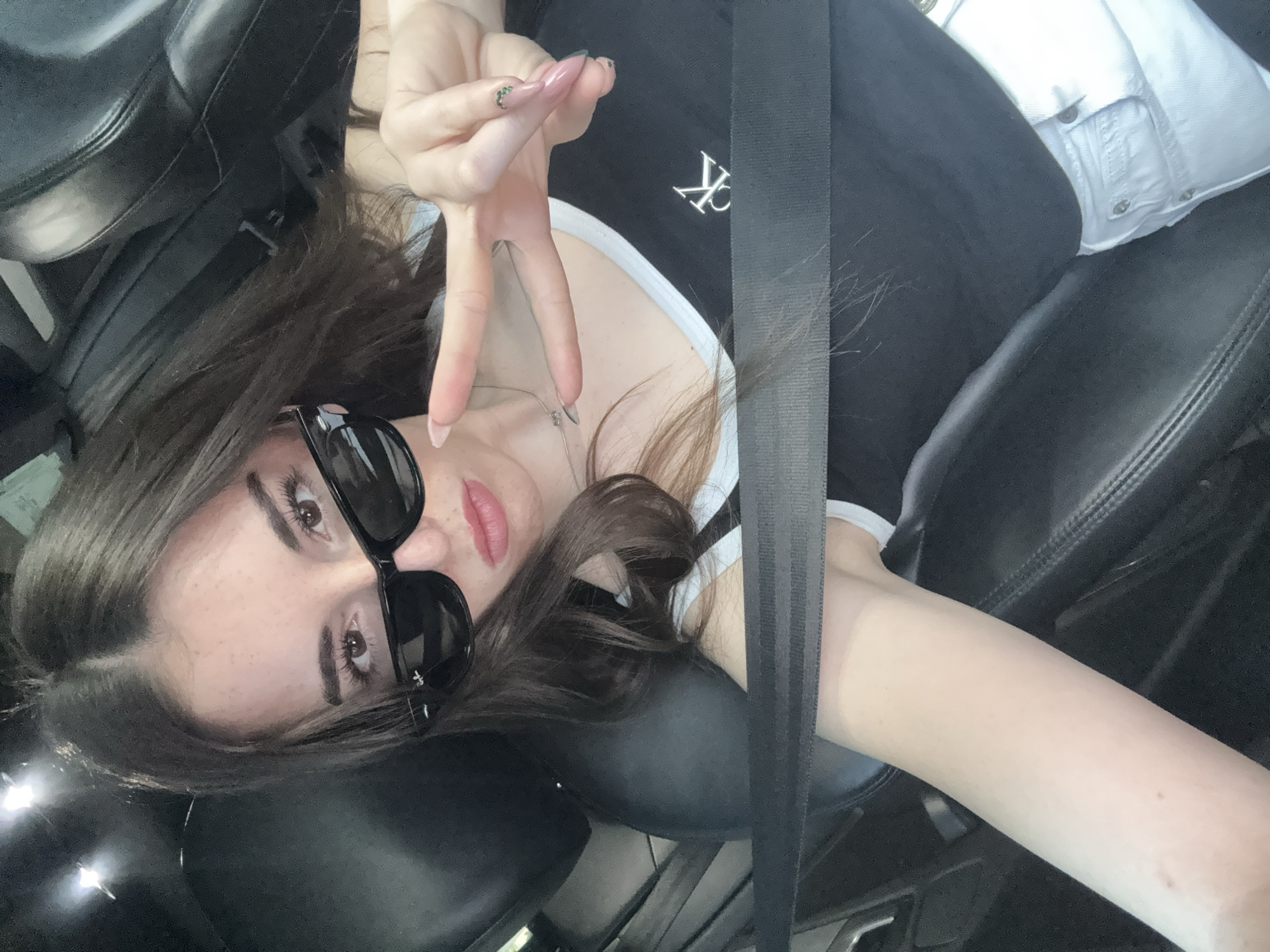 A young woman is seated in a vehicle, wearing a black tank top with white trim and a small, stylized letter "K" logo on the left chest. She is wearing black sunglasses and a seatbelt. Her left hand is raised, displaying a peace sign with her fingers. The background shows black leather seats and interior elements of the car. The image does not provide enough information to determine the exact location within Plovdiv, Bulgaria.