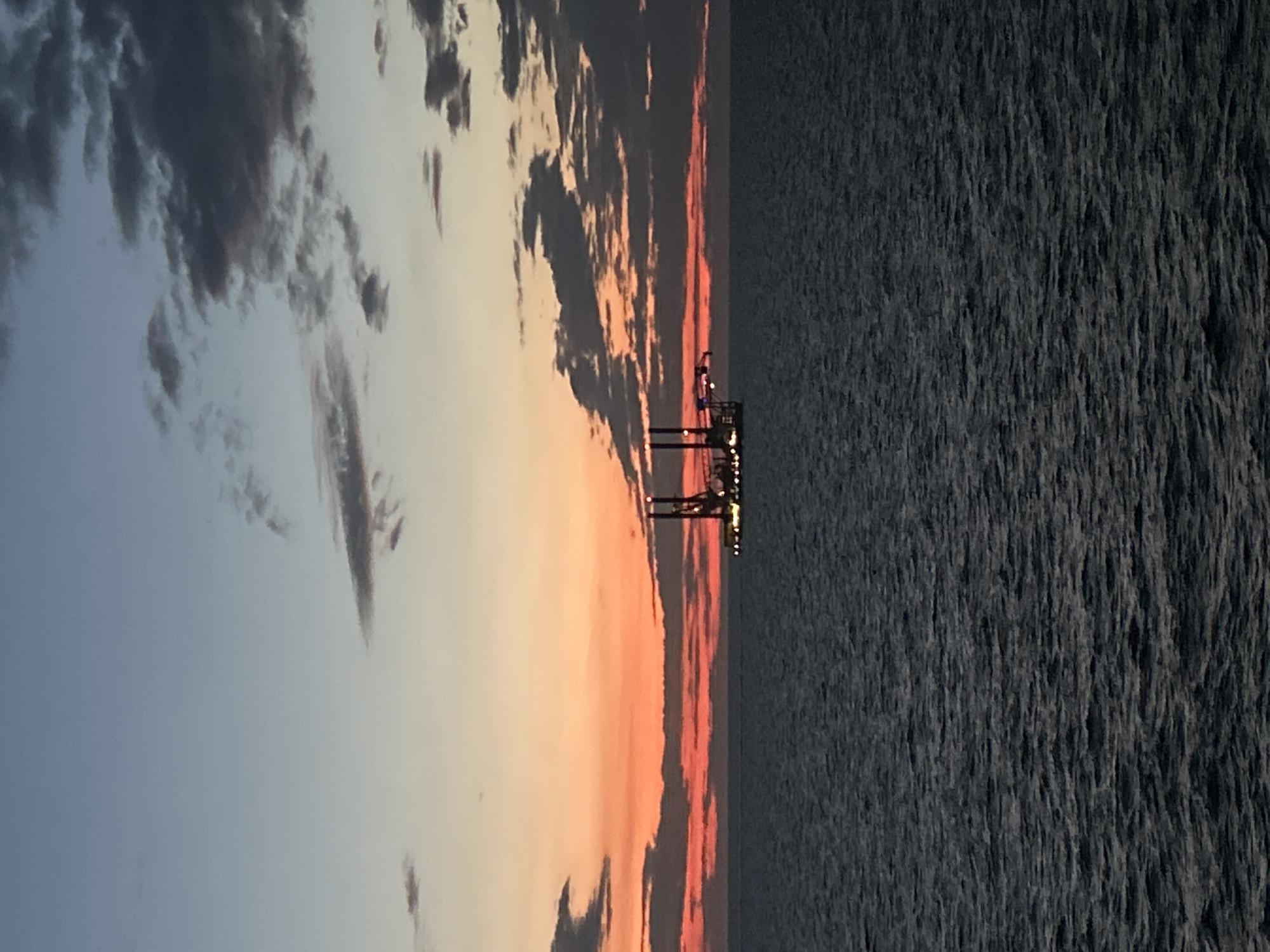 The image depicts an expansive open ocean scene at dusk or dawn, with the location identified as Unknown, Unknown. The sky, occupying approximately two-thirds of the frame, exhibits a distinct color gradient: a pale blue-grey at the zenith transitions downwards into a vibrant band of orange and red hues directly above the horizon, characteristic of twilight. Overcast conditions are present, with scattered dark grey clouds distributed across the upper and middle portions of the sky, particularly dense along the red horizon line.
The ocean, dark and rippled, fills the lower one-third of the image, displaying subtle texture from small waves. Positioned slightly right of the vertical center and resting on the ocean surface is an offshore industrial platform. This structure, appearing as a dark silhouette against the luminous horizon, is illuminated by numerous small, bright yellow-orange lights outlining its perimeter and various components. Prominent vertical structures, likely legs or towers, rise from the platform's deck, which also features horizontal extensions that could be cranes or other equipment. No human figures or overt actions are discernible.