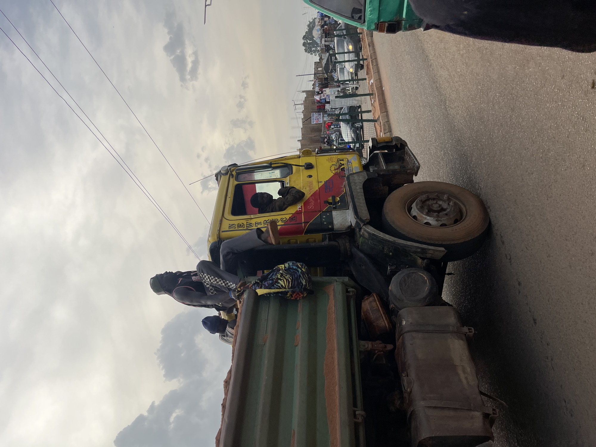 The image depicts a street scene in Bukuru, Nigeria, under an overcast sky. A large yellow dump truck with a green bed filled with reddish-brown dirt is the primary subject. Several individuals are positioned on the truck; one man is sitting on the edge of the truck bed, facing right, wearing a dark tank top, checkered shorts, and a cap. Another person, wearing a blue beanie, is sitting behind him. A third individual is visible inside the truck's cabin, looking out the driver's side window. A colorful patterned bag hangs from the side of the truck. The truck is on a paved road, with other vehicles and buildings visible in the background, suggesting a busy urban environment. A green vehicle is partially visible in the foreground on the right. Power lines are strung across the sky.