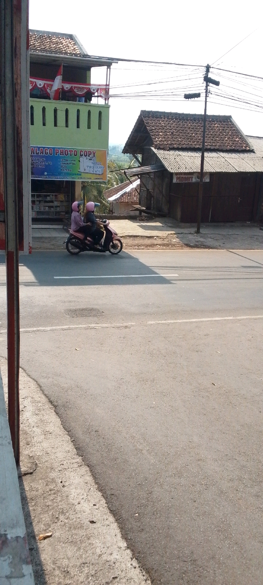 In the city of Cilaku, Indonesia, two individuals, one riding and one pillion, are on a pink motorcycle traveling on an asphalt road. Both are wearing pink helmets. The motorcycle is passing by a light green building with white railings on the second floor, which displays a banner that reads "ALAGO PHOTO COPY" and lists services such as "FOTO DIGITAL", "CETAK FOTO", "LAMINATING", "JILID", and "MATERAI". To the right of the green building is a structure with a tiled roof and dark wooden doors. Overhead, power lines are visible against a bright sky. The road has a white line marking. The foreground shows the edge of a concrete sidewalk and a gray asphalt road surface.