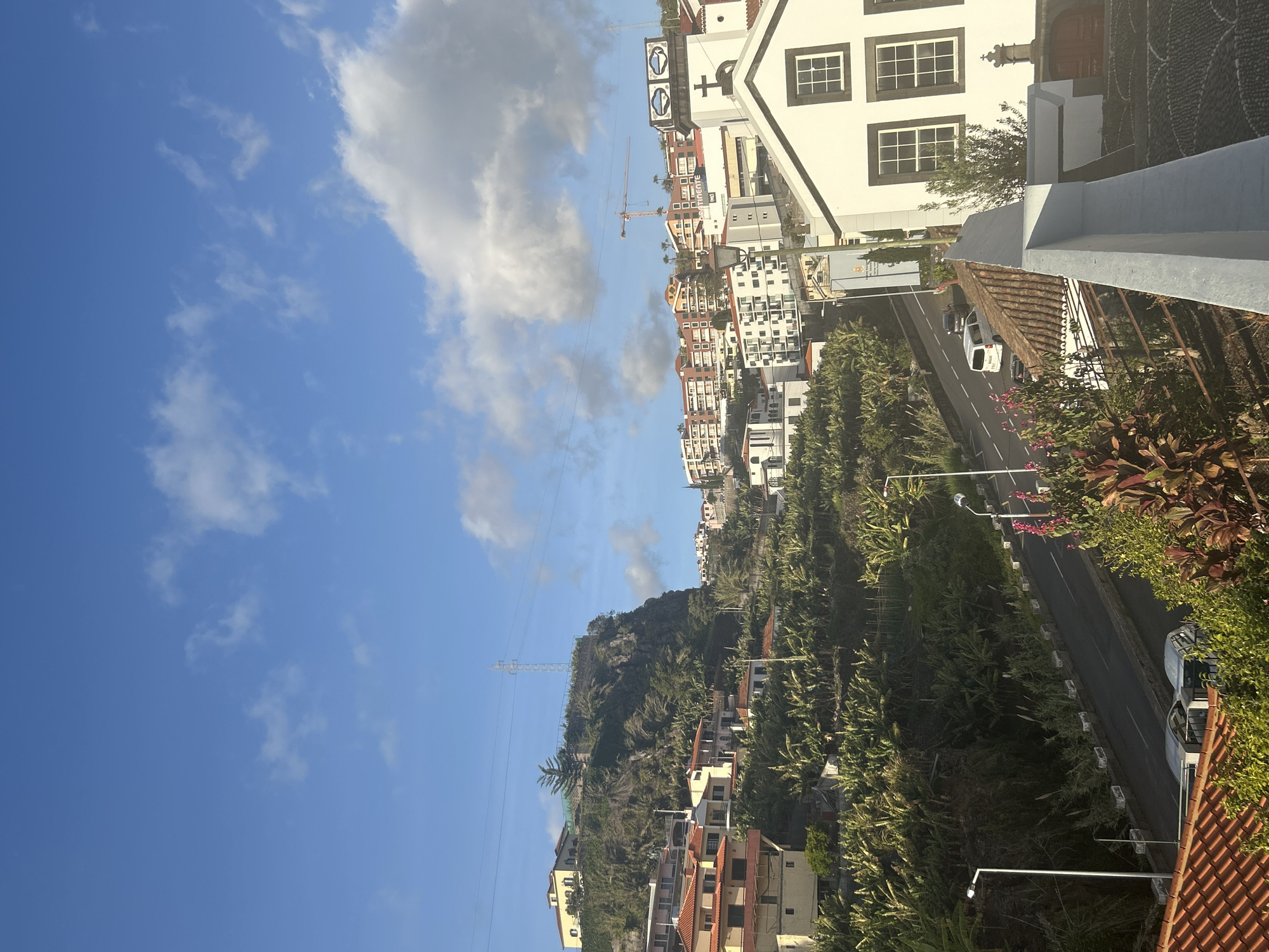 The image shows a hillside scene in Câmara de Lobos, Portugal. A blue sky with scattered clouds is visible. The landscape includes a hill with houses, a road with sparse traffic, and dense vegetation. In the foreground, there is a building with white and black accents. In the distance, a construction crane can be seen among buildings. Power lines are also visible extending across the sky.
