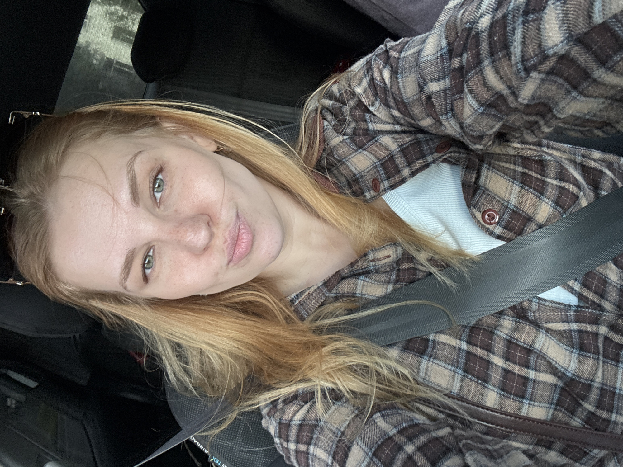 The image is a selfie taken inside a vehicle. A young woman with long, blonde hair is wearing a plaid shirt and a seatbelt. The vehicle appears to be in motion, with the background showing blurred scenery. The specific location of Podosenikha, Russia is not discernible from the image.