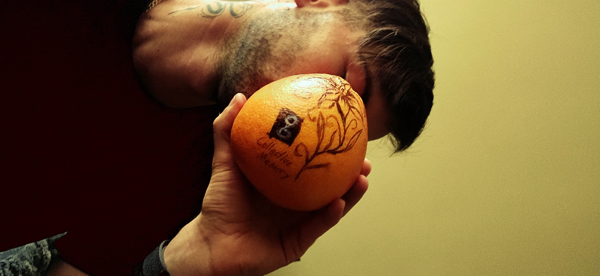A man is holding an orange in his right hand. The orange has drawings on it, including a square with a symbol resembling "8" inside, the words "Collective Memory" written below it, and a floral design. The man has a tattoo on his neck. He is wearing a red shirt and a denim jacket. The background is a solid yellow wall. The photo does not provide enough information to determine the location in the city of Plovdiv, Bulgaria.