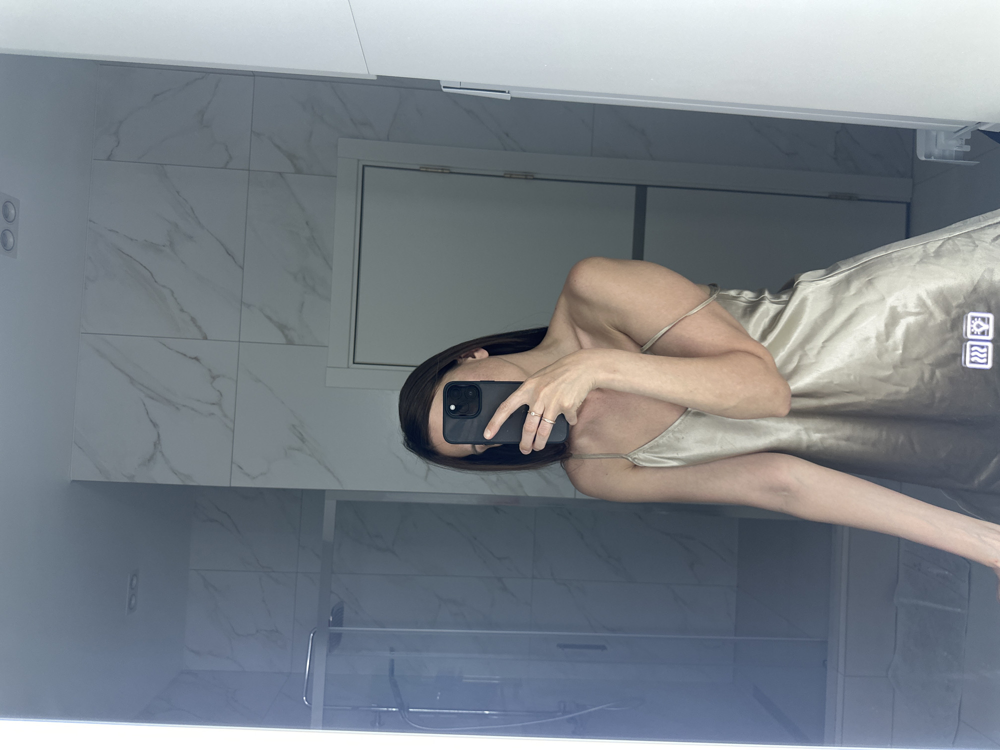 A person is taking a selfie in a bathroom. The person has dark hair and is wearing a light-colored satin-like slip dress. The bathroom has white marble-patterned tiles on the walls. There is a shower stall with a glass door and a white cabinet above the toilet. The reflection in the mirror suggests the bathroom is in Прикамский, Russia.