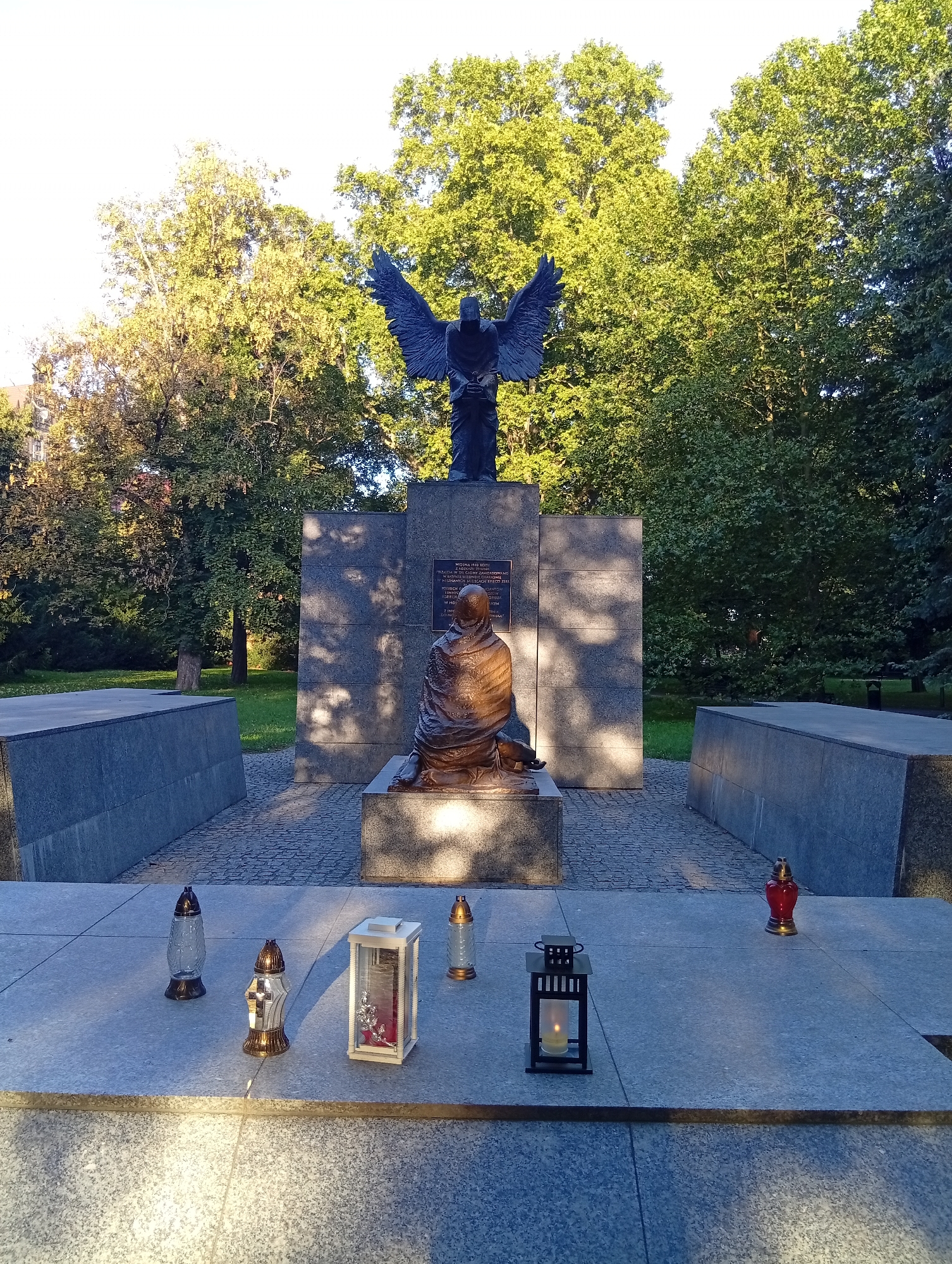 The image shows a monument in Wrocław, Poland, featuring a bronze angel statue with outstretched wings standing atop a stone pedestal. In front of the pedestal, a bronze statue of a seated, cloaked figure is positioned on a lower base. Behind these figures is a large, grey stone monument with inscribed text. The monument is located in a park-like setting with mature trees in the background. In the foreground, on a grey stone platform, several memorial candles are placed: two tall, glass-encased candles with lids (one with a cross emblem), a white rectangular lantern-style candle holder with a lit candle, and a red, flame-shaped candle. The scene is illuminated by natural sunlight, casting shadows on the monument.
