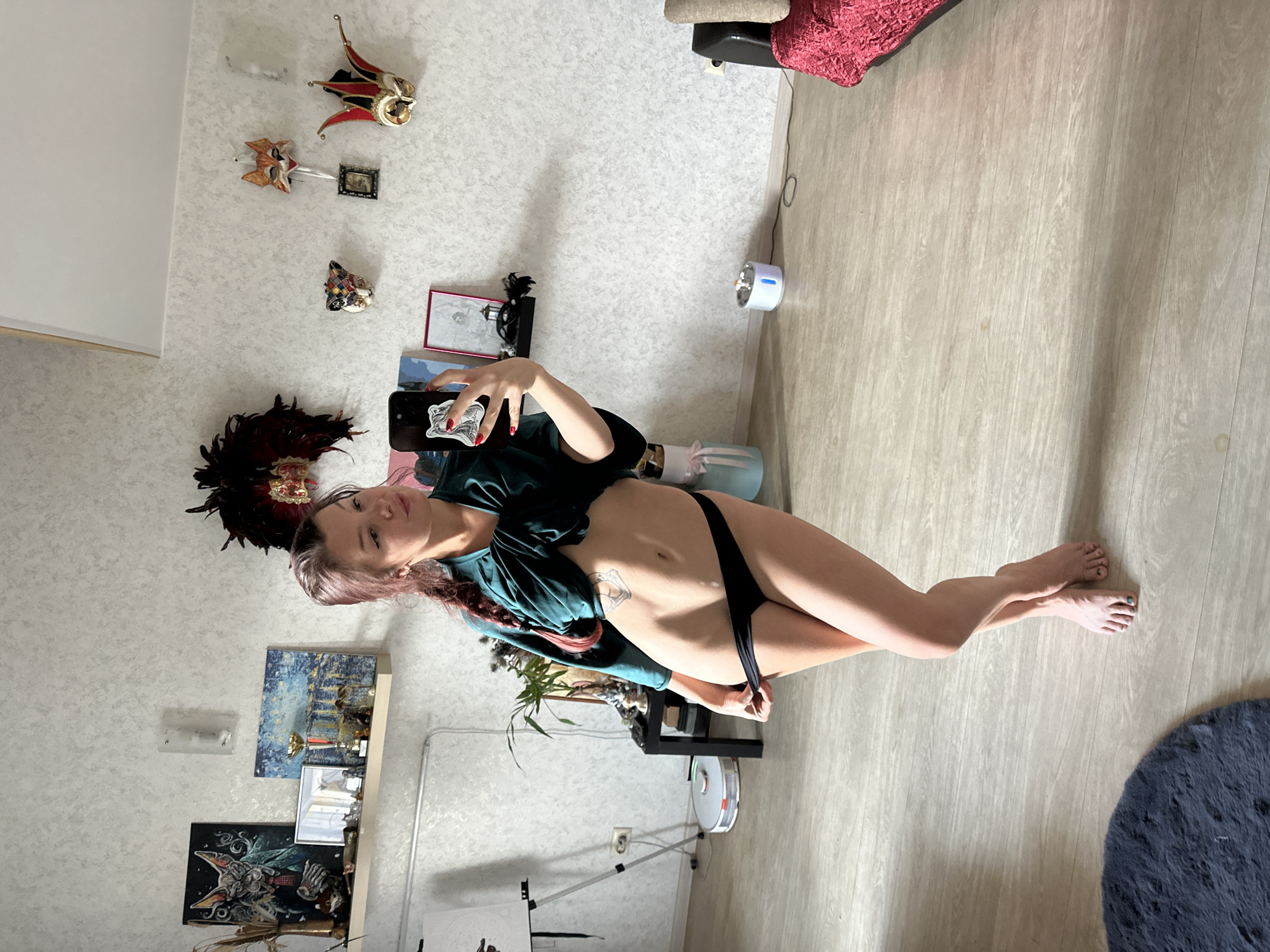 A woman is taking a selfie in a room with light-colored textured walls and light wood-plank flooring. She is wearing a dark green off-the-shoulder top and black underwear. Her hair is long and has pinkish streaks. She is positioned in the center of the frame, looking towards the camera.
Various decorative items are visible in the room. On the wall above her, there are several Venetian-style masks in red, gold, and white. Below these, there are framed pictures and other wall hangings. To the left, a shelf holds more artwork, trophies, and decorative objects. A tall, dark feather headdress is also present on a shelf near the masks.
In the background, a white and blue air purifier or humidifier is on the floor near a dark cabinet. A robotic vacuum cleaner is also visible on the floor. A tripod with a canvas on it stands in the lower part of the image, suggesting an art setup. A dark blue furry rug is partially visible in the lower right corner. The lighting suggests it is daytime, with light coming from the left. The overall mood is casual and personal. There is no visible text in the image.