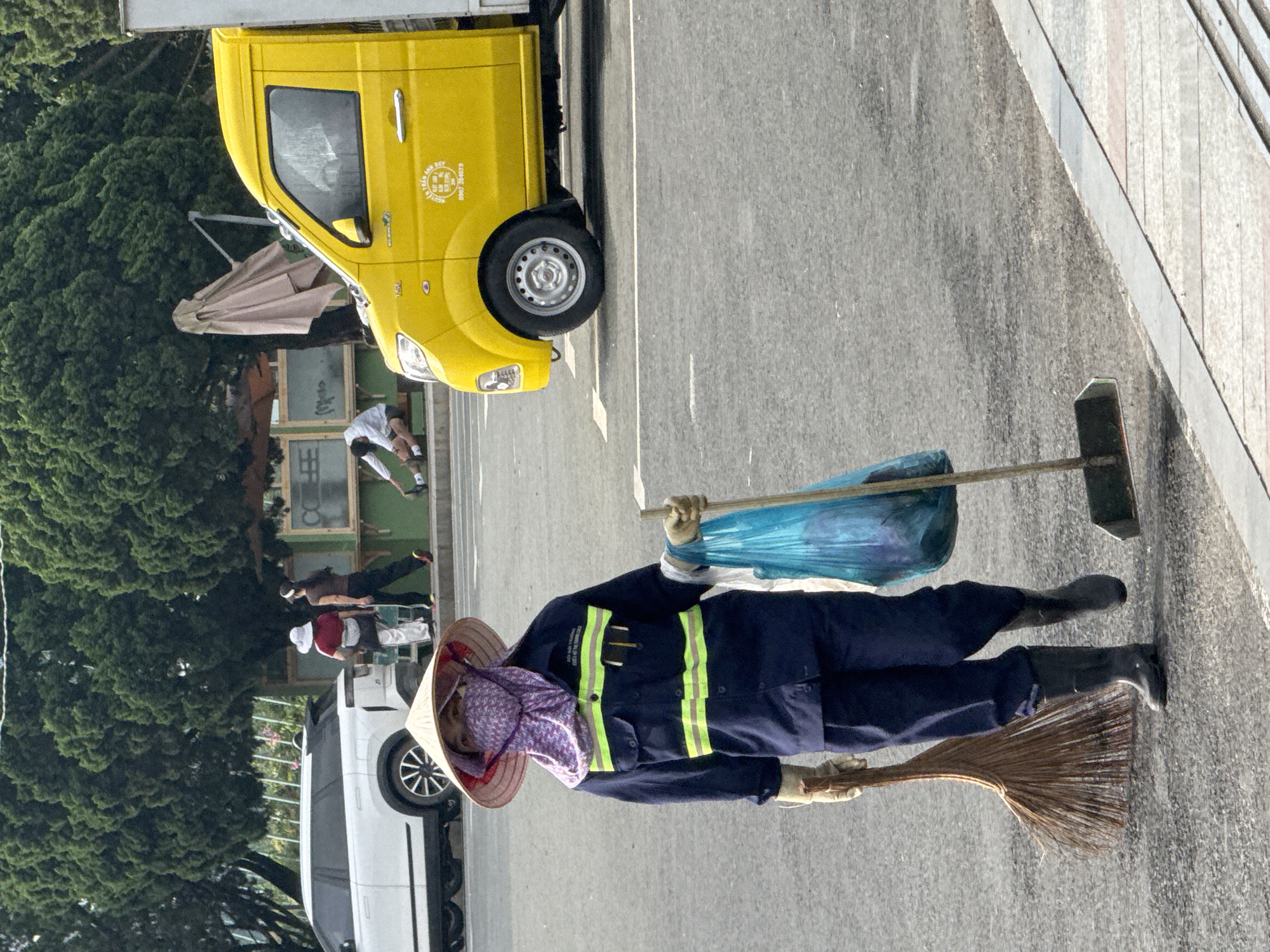 The image shows a street scene in Da Lat, Vietnam. In the foreground, a person wearing a dark blue uniform with reflective yellow stripes, a conical hat, and a face covering is walking across the asphalt. They are carrying a broom in their left hand and a blue plastic bag filled with trash in their right hand, holding a broom handle. To the right, a bright yellow truck is parked on the street. In the background, several people are visible. Two individuals stand near a white SUV. Further back, another person is sitting on a bench, and another person is standing. Tall green trees line the background, with some structures and umbrellas also present. The ground is paved with asphalt and has white line markings.