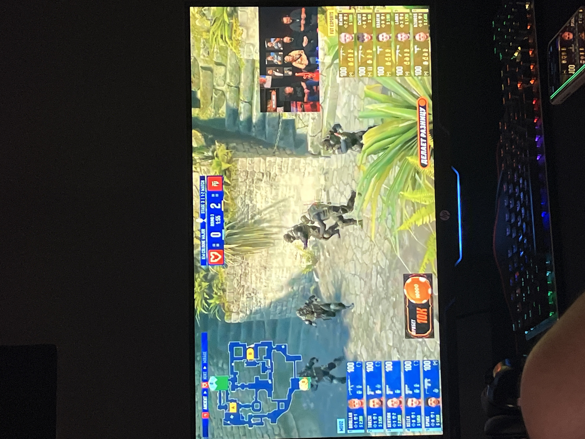 The image shows a computer screen displaying a Counter-Strike 2 esports match. The game is being played on the map "Ancient." The score is 0-2 in favor of the team on the right. The players' names and stats are visible on the left and right sides of the screen. The game is part of the "IEM Cologne Major." The background of the game shows a digital representation of an ancient, stone-walled environment with lush greenery. On the right side of the screen, a broadcast of the casters is visible, with several individuals seated and watching the game. The computer is connected to a keyboard with RGB lighting, and a smartphone is placed in the foreground. The scene appears to be indoors, likely a gaming setup. There is no indication of the specific location being Chaikovsky, Russia.