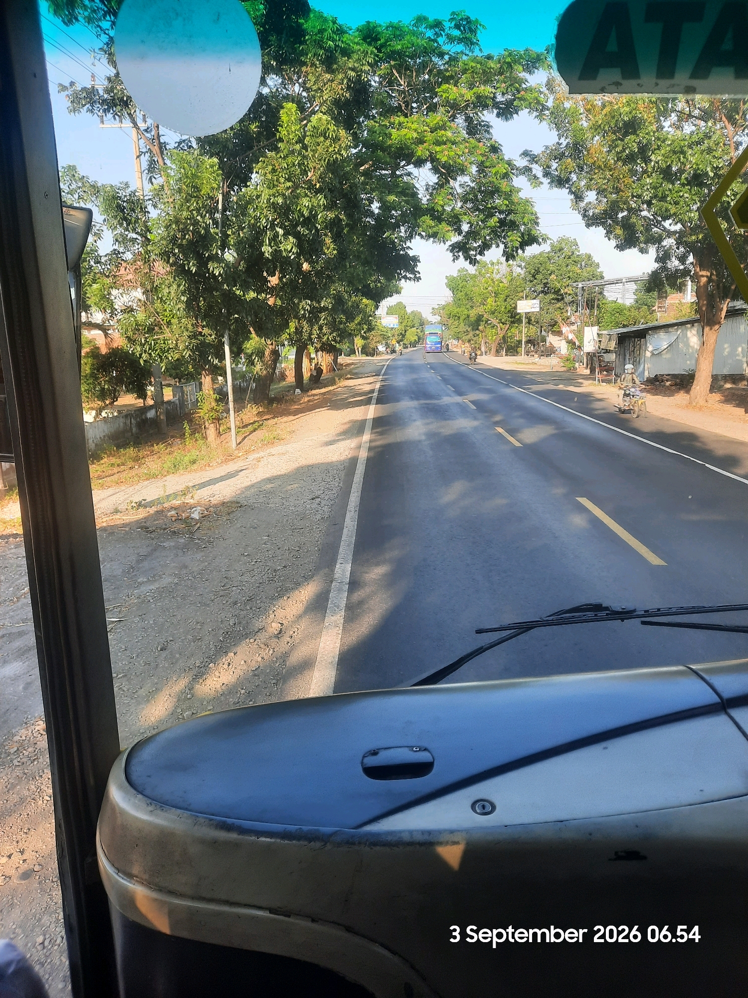 The image depicts a view from inside a vehicle, looking out at a road in Sukokerto, Indonesia. A dashboard is visible in the foreground, with a windshield and wipers. The road stretches into the distance, lined with trees on both sides. A blue bus and a motorcycle are seen on the road. The sky is clear and blue. The date and time stamp on the image indicate "3 September 2026 06.54". The overall scene suggests a sunny morning on a suburban or rural road.