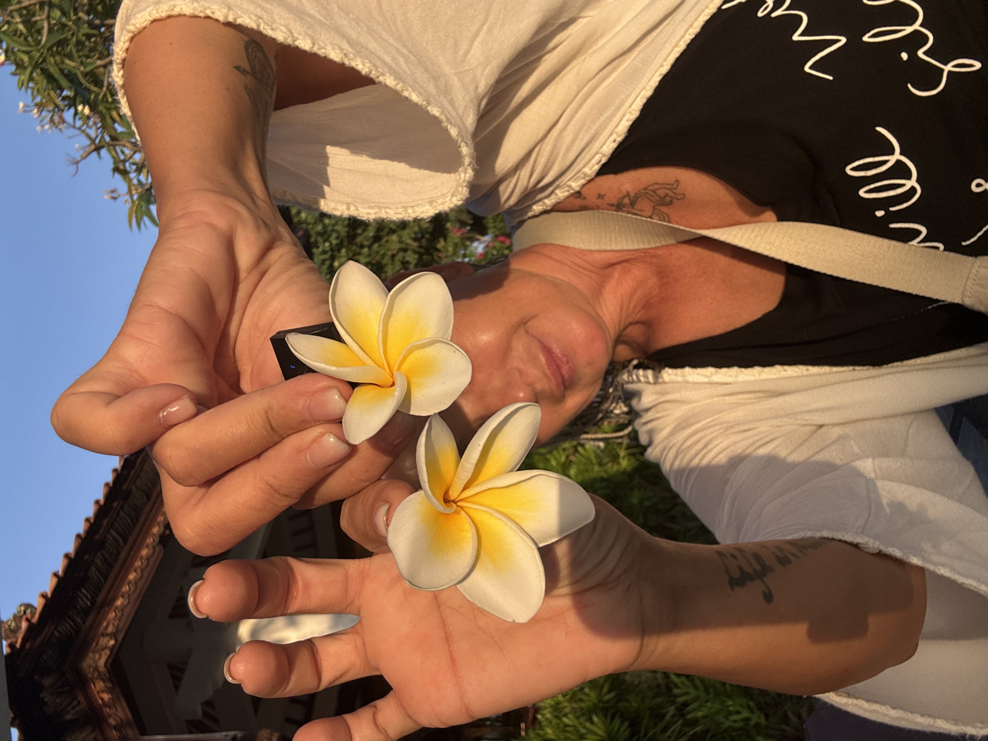 In Kuta Selatan, Indonesia, a woman holds two white and yellow plumeria flowers, one partially obscuring her left eye, while her right hand holds another flower near her face. She wears a white, open-front cardigan over a black t-shirt with white cursive text. A light-colored strap crosses her chest. Tattoos are visible on her upper left arm and near her collarbone. The background features a traditional Balinese building with a thatched roof, lush green foliage, and a clear blue sky. The scene is illuminated by the warm glow of late afternoon sunlight.