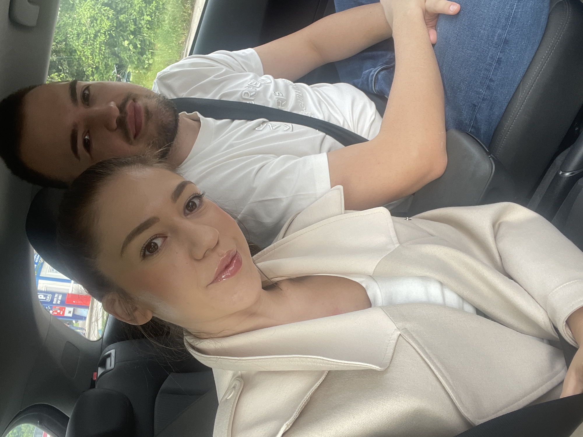 Inside a car, a man and a woman pose for a photograph, likely a selfie. The image is oriented sideways, with the subjects appearing to lie down, but they are seated upright in the vehicle. The woman is in the foreground, facing the camera with a neutral expression and glossy lips. She wears a cream or beige overcoat over a white top. Her hair is pulled back, and her eyes are dark brown.
Behind and to her right is a man with a beard and dark hair, also looking at the camera with a neutral gaze. He is wearing a white t-shirt with embossed or printed text, including "KAP" and parts of "R RIE". He is dressed in blue jeans. Both individuals are wearing black seatbelts. The man's arm rests on his leg, and his hand is clasped with the woman's hand, suggesting a close relationship.
The setting is the interior of a car, with black seats and interior trim visible. Through the car window in the background, green foliage is seen, indicating an outdoor, daytime setting. Soft, diffused light suggests an overcast or partly cloudy sky. To the left of the woman's head, outside the car, a structure with blue and red elements is visible, possibly a gas station or commercial building. Partial text, including "R2" and "PET", can be discerned on this structure. The scene is located in Sofia, Bulgaria.