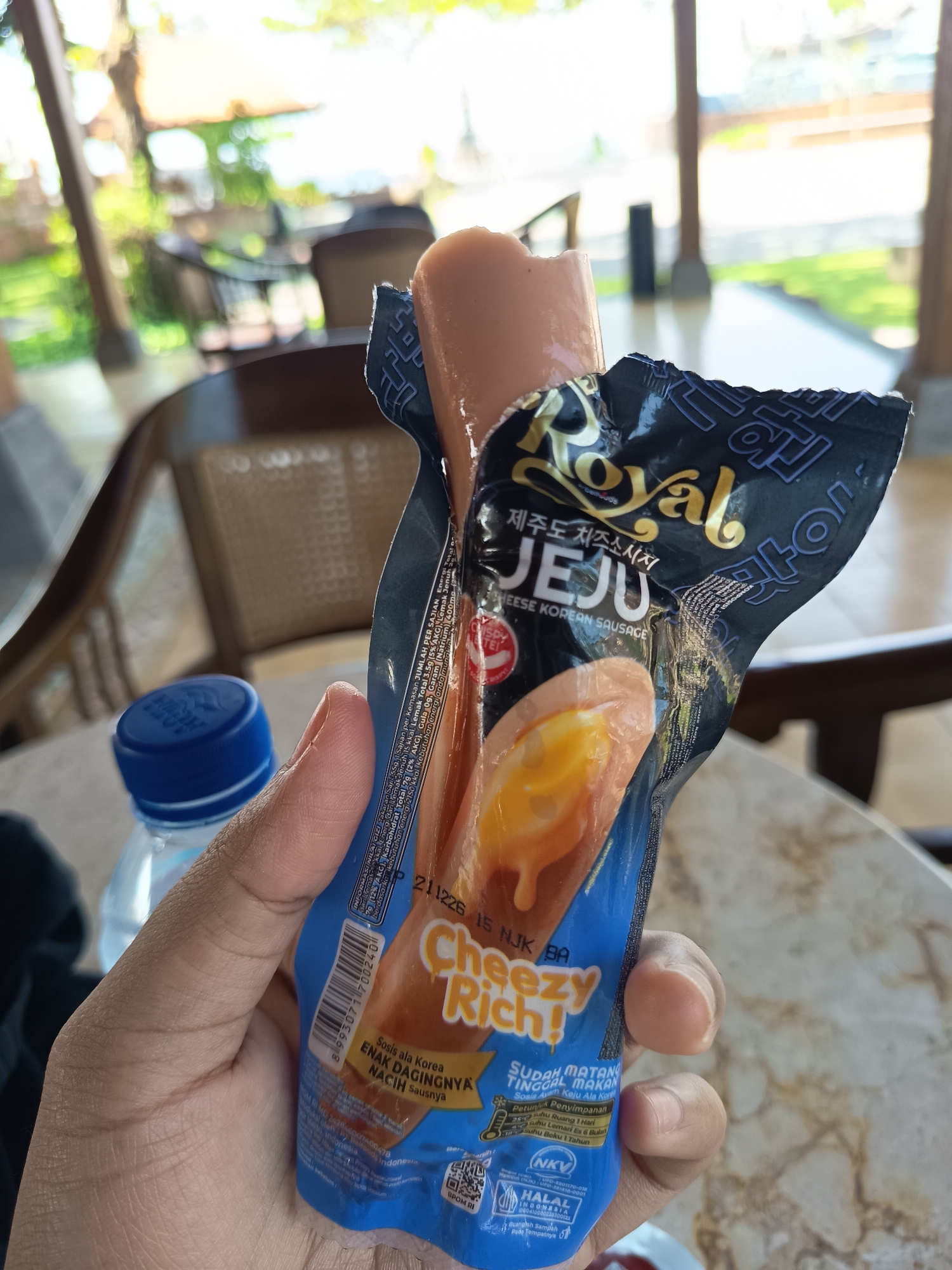 A hand holds an opened package of "Royal Jeju Cheese Korean Sausage." The sausage is a light brown color with a visible yellow cheese filling peeking out. The packaging is primarily black with gold and white text, featuring the brand name "Royal" in a stylized font and "JEJU CHEESE KOREAN SAUSAGE" in English. Below this, it says "Cheezy Rich!" in large, playful lettering. The packaging also displays nutritional information and certifications. In the background, a blue water bottle cap is visible. The scene appears to be outdoors, with blurred elements of trees and buildings suggesting a tropical setting, possibly a resort or restaurant patio in Denpasar, Indonesia, characterized by the open-air architecture and lush greenery.