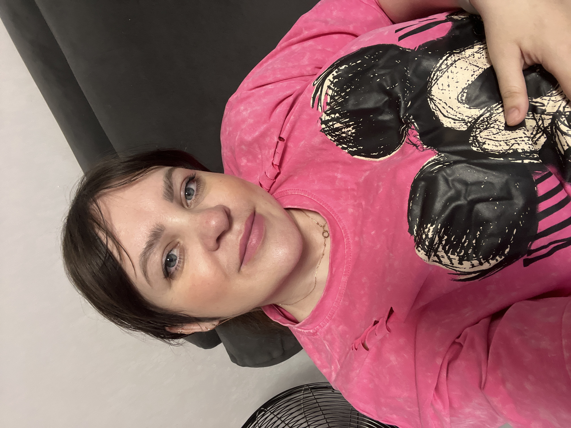 An indoor selfie, tilted to the left, features a young to middle-aged woman lying in a relaxed pose. She has fair skin, dark brown hair, and bright blue eyes, with a gentle, soft smile on her lips. Her makeup is subtle, accentuating her eyes. She is wearing a vibrant, distressed pink long-sleeved shirt or sweatshirt with a prominent black and white graphic resembling a stylized cartoon character, possibly Mickey Mouse. One of her hands rests casually on this graphic. A delicate gold-colored chain necklace is visible around her neck. The background includes a dark grey surface, possibly a sofa or headboard, and a lighter grey or white wall. A black metal fan grill is partially visible in the bottom right corner. The lighting suggests an indoor setting, likely with artificial light. The woman appears calm and comfortable. While the image itself provides no location cues, it is set in Perm, Russia. There is no visible text in the media.