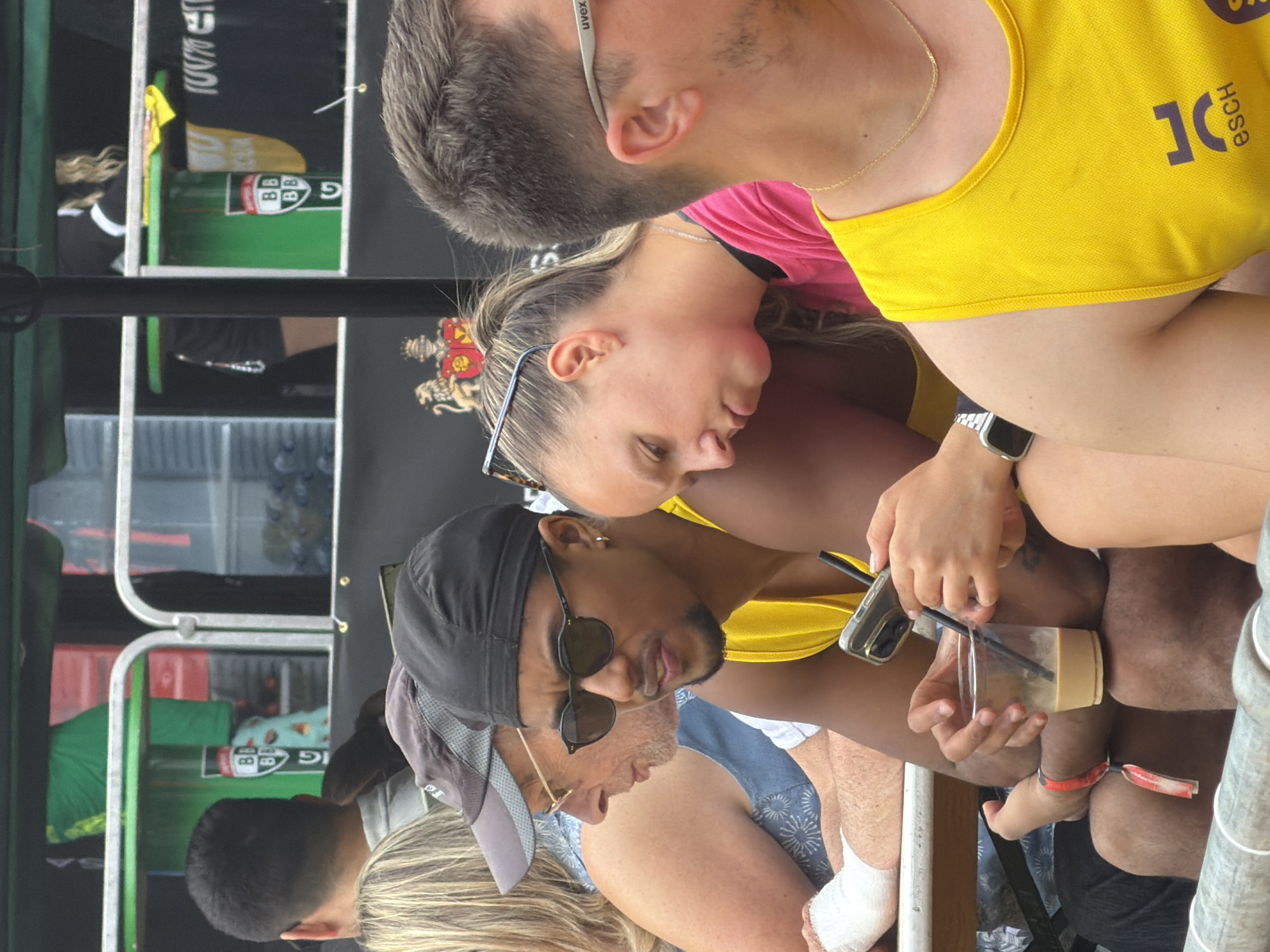 The image depicts a group of people seated outdoors, possibly at a stadium or event venue in Esch-sur-Alzette, Luxembourg, given the presence of the "esch" logo on the yellow tank top. In the foreground, a man wearing a black durag and sunglasses is holding a phone and a drink with a straw. To his left, a woman with blonde hair pulled back in a ponytail is looking down at her phone. To the right of the man, another person wearing a yellow tank top with the "esch" logo is visible. In the background, there are glimpses of what appears to be a food or drink stall with green elements and some signage. The overall setting suggests a public gathering or sporting event.