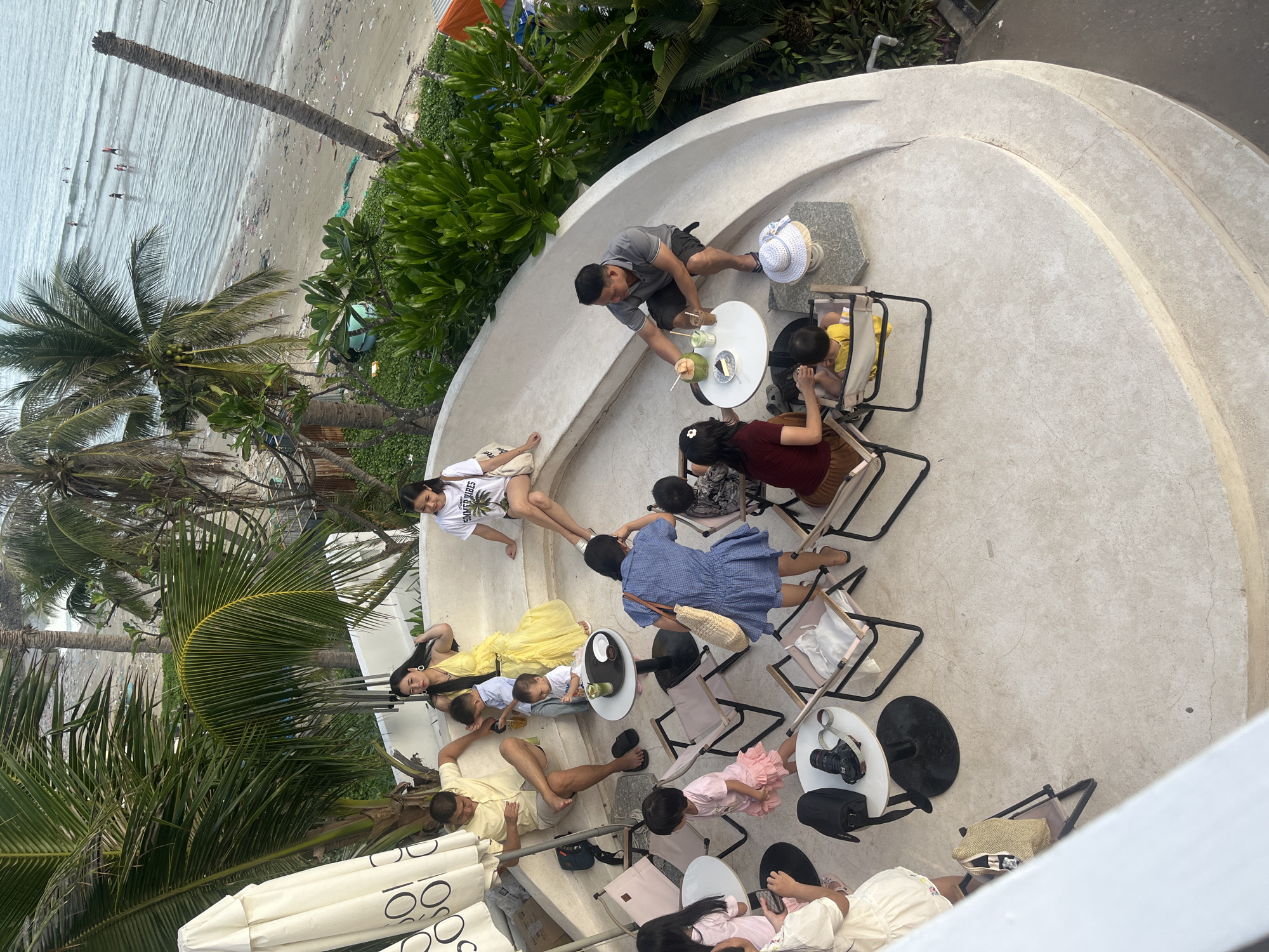 An aerial view captures a scene at a beachfront cafe in Mũi Né, Vietnam, with a partial view of the ocean and beach in the background. The cafe's outdoor seating area is constructed from light-colored concrete, featuring a curved seating ledge and circular platforms for tables and chairs.
Several groups of people are present. In the background, a family is seated on the curved ledge. A man in a yellow short-sleeved shirt and shorts sits with his legs crossed, a child in a blue shirt next to him. A woman in a bright yellow dress is seated with a baby on her lap. Another woman in a white t-shirt with graphics is seated on the far right of the ledge.
In the foreground, another group is gathered around a small, round table. A man in a gray polo shirt and dark shorts holds a coconut drink and a straw. A woman in a dark red top and brown skirt sits facing him. A young child in a yellow outfit is in a portable high chair.
Scattered throughout the seating area are several portable chairs with light-colored fabric. Small, round tables are positioned near the chairs. Objects on the tables include drinks, plates, a straw, a hat, and a camera bag with a camera on top.
The environment includes palm trees with lush green fronds, and further back, the ocean with a sandy beach littered with debris. The sky is overcast, and the overall lighting suggests late afternoon.