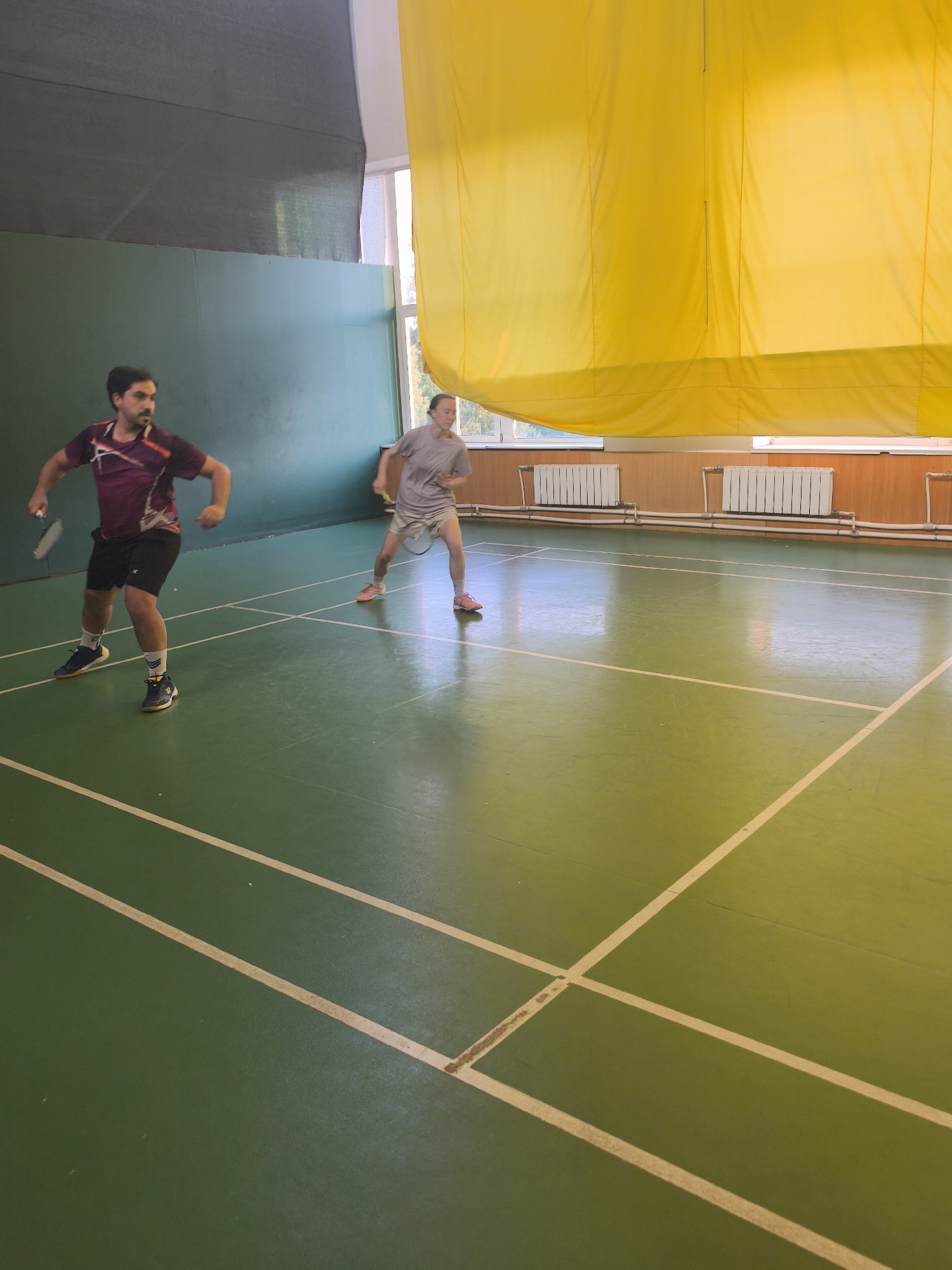 This image captures an indoor sports facility located in Saint Petersburg, Russia. Two individuals are actively playing badminton on a green court delineated by white lines. On the left, a male subject with dark hair and a beard, wearing a maroon and white athletic shirt, black shorts, and blue and yellow athletic shoes, stands poised with a badminton racket in his right hand. On the right, a female subject with light hair tied back, dressed in a grey t-shirt, light beige shorts, and pink athletic shoes, similarly holds a badminton racket in her right hand, exhibiting a ready stance. The background wall on the left is a dark teal color, while the right wall features light wooden paneling below several large windows, through which outdoor foliage is visible. White radiators and associated piping are mounted along the wooden paneling. A large, bright yellow fabric panel hangs from the ceiling across the upper right section of the room, partially covering the windows. The green floor shows visible signs of wear and dirt along the white court lines.