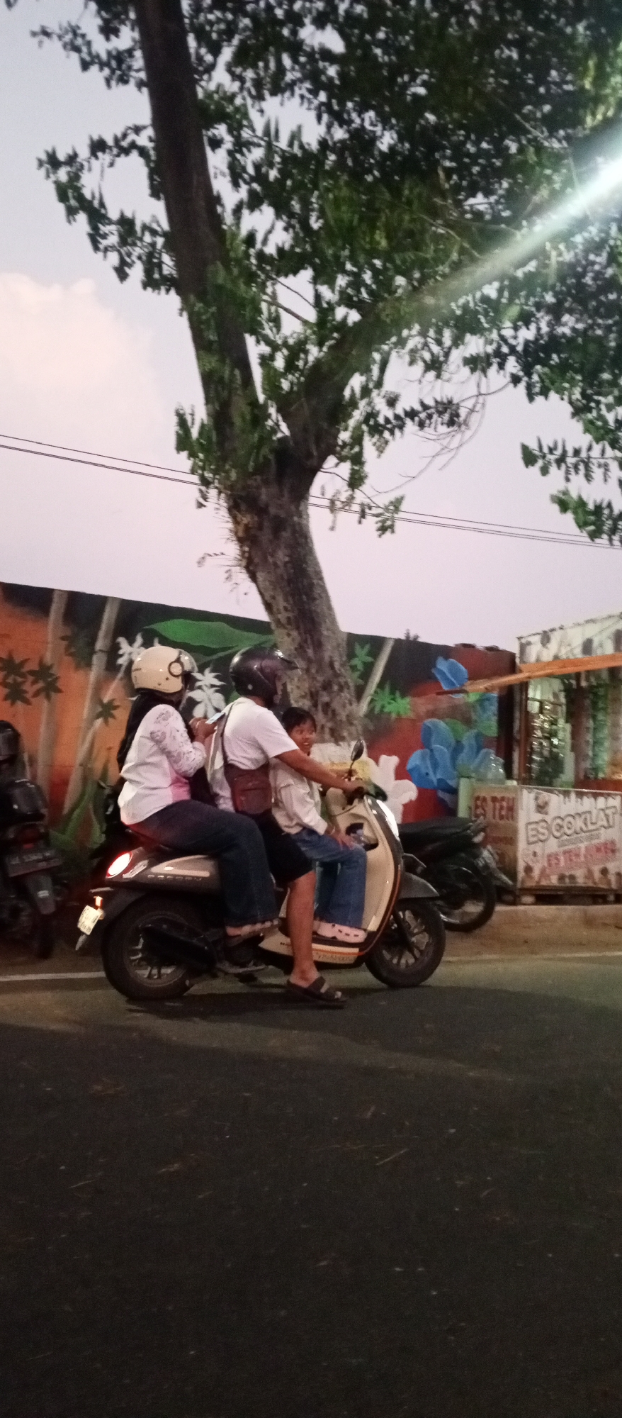A family of three is riding a scooter at dusk. The father is driving, the mother is in the middle, and their child is in front. They are all wearing helmets. The scooter is a cream-colored scooter. They are on a street in Madiun, Indonesia, near a mural of a tree and flowers. There is a stand selling drinks in the background. The sky is a pale purple and the tree has green leaves.