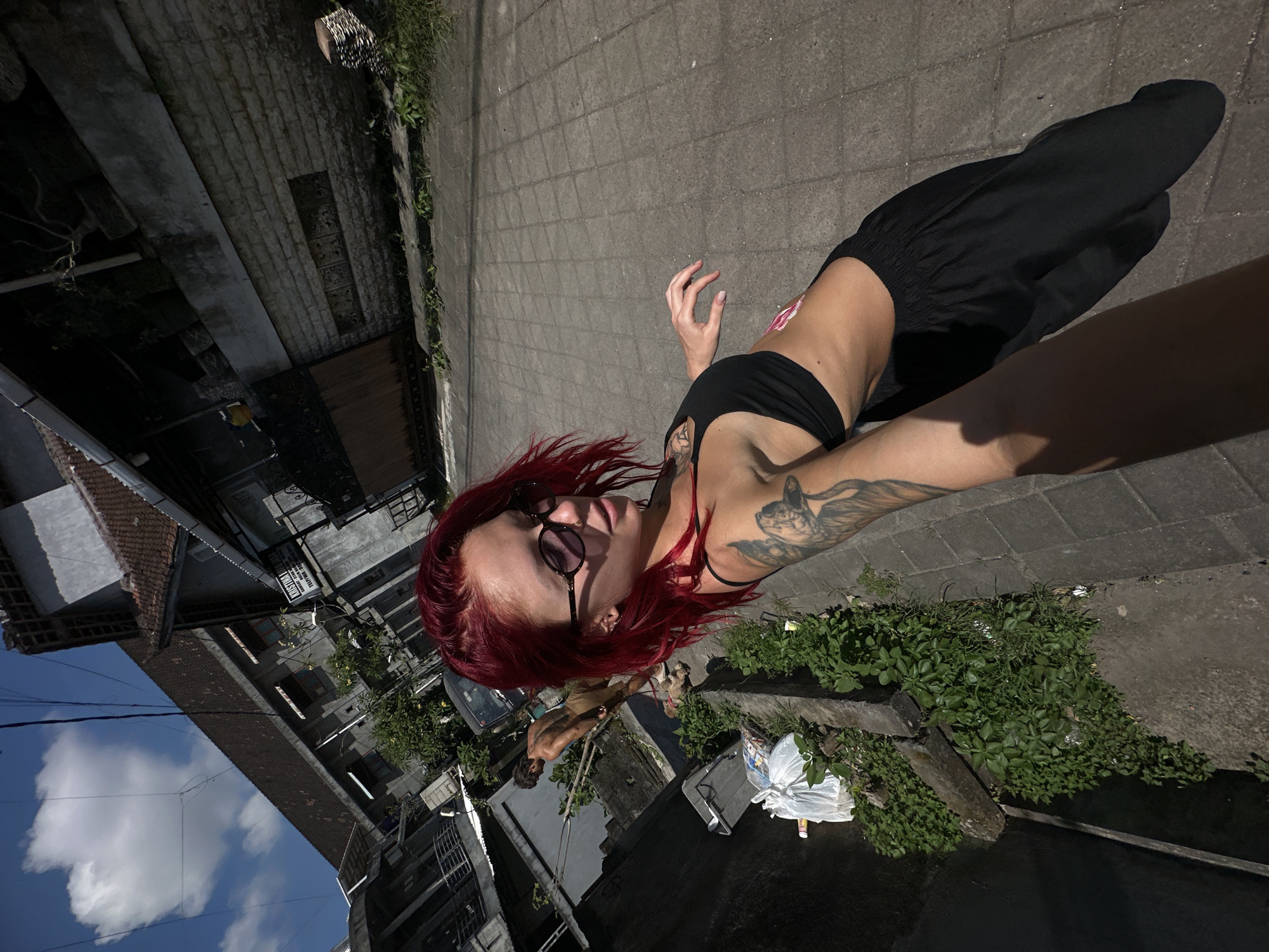 This image captures a vertical selfie taken in an urban environment in North Kuta, Indonesia, under bright daylight. The main subject is a person with vibrant red hair and dark sunglasses, wearing a black crop top and black loose-fitting pants. They are looking directly at the camera with a playful expression, their tongue slightly extended. Tattoos are visible on both arms, including a prominent feline head on their left shoulder/arm.
The person's right arm is outstretched, holding the camera, with their body slightly turned. In the background, a paved walkway extends into the distance, bordered by various buildings and green vegetation. Another person, a shirtless man with dark skin, is visible further back, walking a small white dog on a leash. The buildings showcase diverse architecture, including structures with tiled roofs and others with concrete walls, interspersed with lush green bushes and vines.
The sky is a clear blue with scattered white clouds, and overhead power lines are visible. Strong shadows suggest it is midday or early afternoon. A partially visible sign on a building in the background reads "KRISTINA" and "TEBING," likely indicating a local business such as a homestay. A car is also parked in the background. The scene conveys a casual, everyday moment in a bustling Indonesian street.