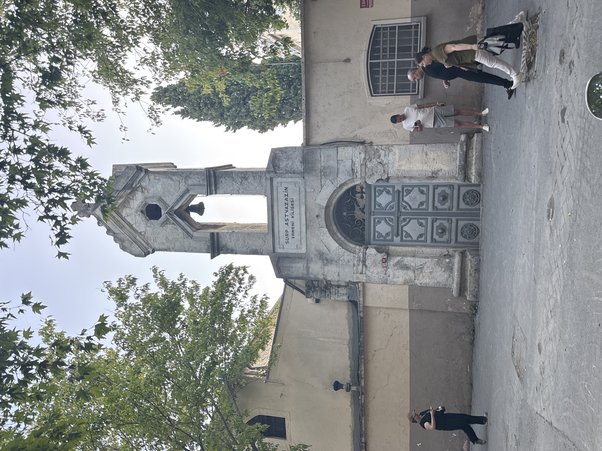 The image shows a street scene in Bakırköy, Turkey, featuring the Surp Astvazazin Armenian Church. A bell tower is prominent in the center of the frame, with an inscription above the arched entrance that reads "SURP ASTVAZAZIN ERMENI KILISESI." The church building is to the left, painted in beige with a darker brown band near the bottom. Several people are present: a woman in black walks from left to right, looking at her phone; a man in a white t-shirt and shorts stands looking at his phone; and a couple walks from right to left. The street is paved with cobblestones and asphalt, with some patches of rougher material and a few small holes. Trees with green leaves are visible in the foreground and background, and the sky is overcast. A red circular sign with the number "30" is visible on the wall to the right of the church entrance.