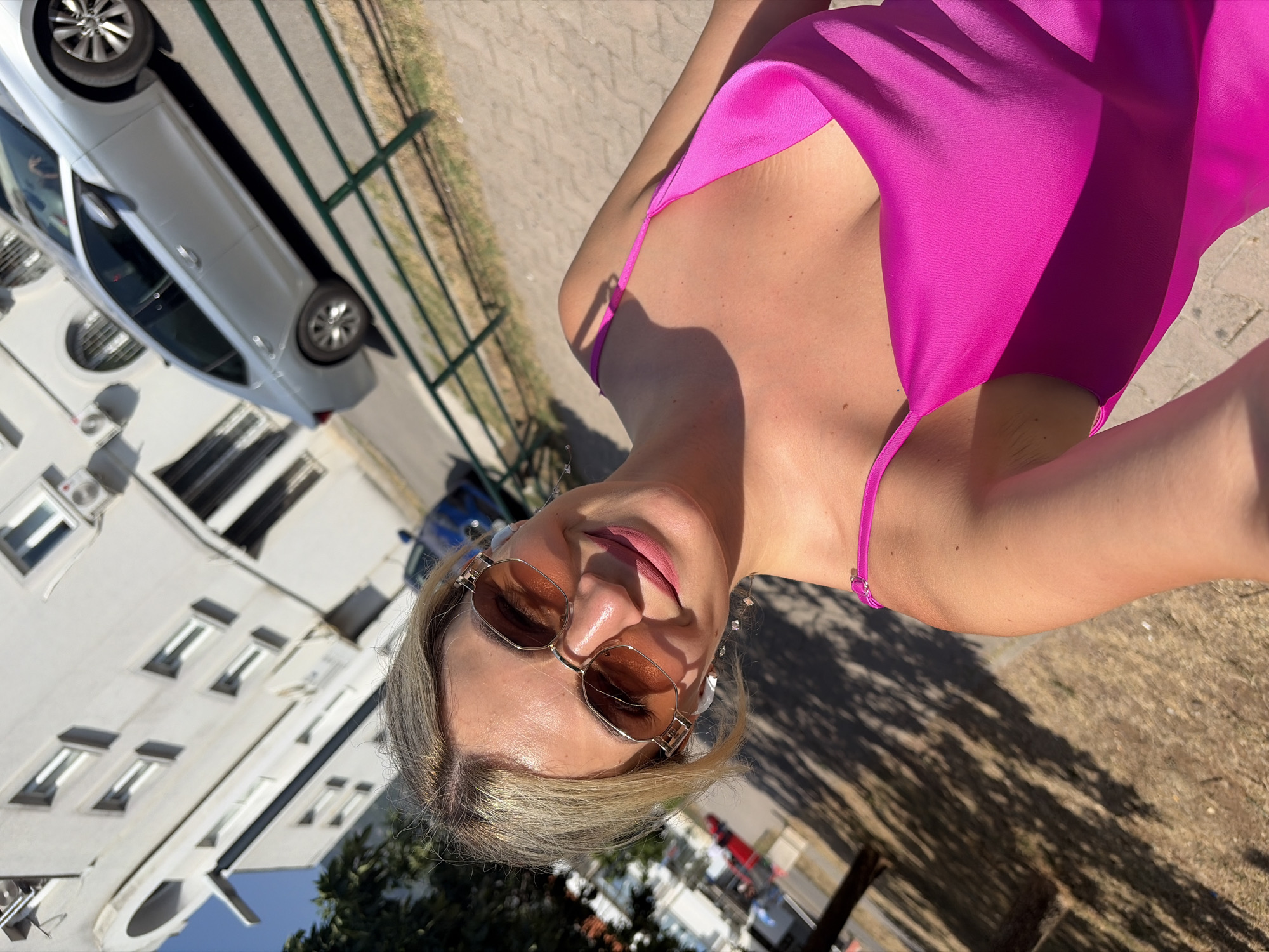 A blonde woman with sunglasses and pink lipstick takes a selfie outdoors in Budva, Montenegro. She is wearing a bright pink slip dress. Behind her, a silver sedan is parked in front of a white building. The ground is paved with light-colored stones, and there are trees and shadows visible. A green metal fence runs along the right side of the image. The sky is a clear blue.