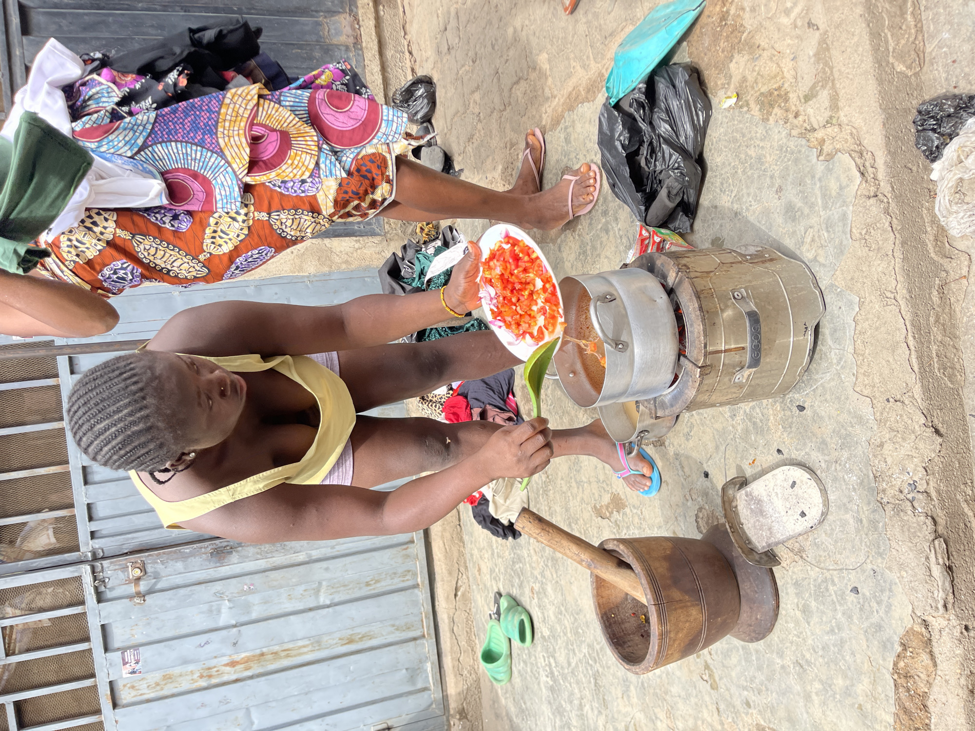 This image depicts an outdoor cooking scene in Nyanya, Nigeria, under bright daylight. Two women are present, engaged in domestic activities.
In the foreground, a woman with neatly braided hair, dressed in a yellow tank top and light-colored undergarments, is actively preparing food. She holds a white plate piled with chopped red tomatoes and onions, poised to add them to a large metal pot resting on a charcoal stove or similar outdoor cooking fire. Her other hand holds a green plastic spoon, suggesting she is also stirring the contents of the pot.
Behind her, partially visible, another woman sits or kneels on the ground, wearing a vibrant, multicolored patterned wrapper or dress and pink flip-flops. Next to her is a pile of clothes, including items with similar colorful patterns.
The cooking setup is situated on a concrete and dirt ground. Nearby, a large wooden mortar with a wooden pestle leaning inside it is visible, indicating traditional food preparation. A pair of green slides or slippers are placed on the ground close to the mortar. Several black plastic bags are scattered around the area, and a sturdy metal gate forms the backdrop to the left. The cooking apparatus has the word "ECOO" visible on its handle.
The scene captures a candid moment of daily life, highlighting traditional cooking methods and the communal nature of household chores in an outdoor setting. The women appear focused on their tasks, reflecting a functional and practical environment.